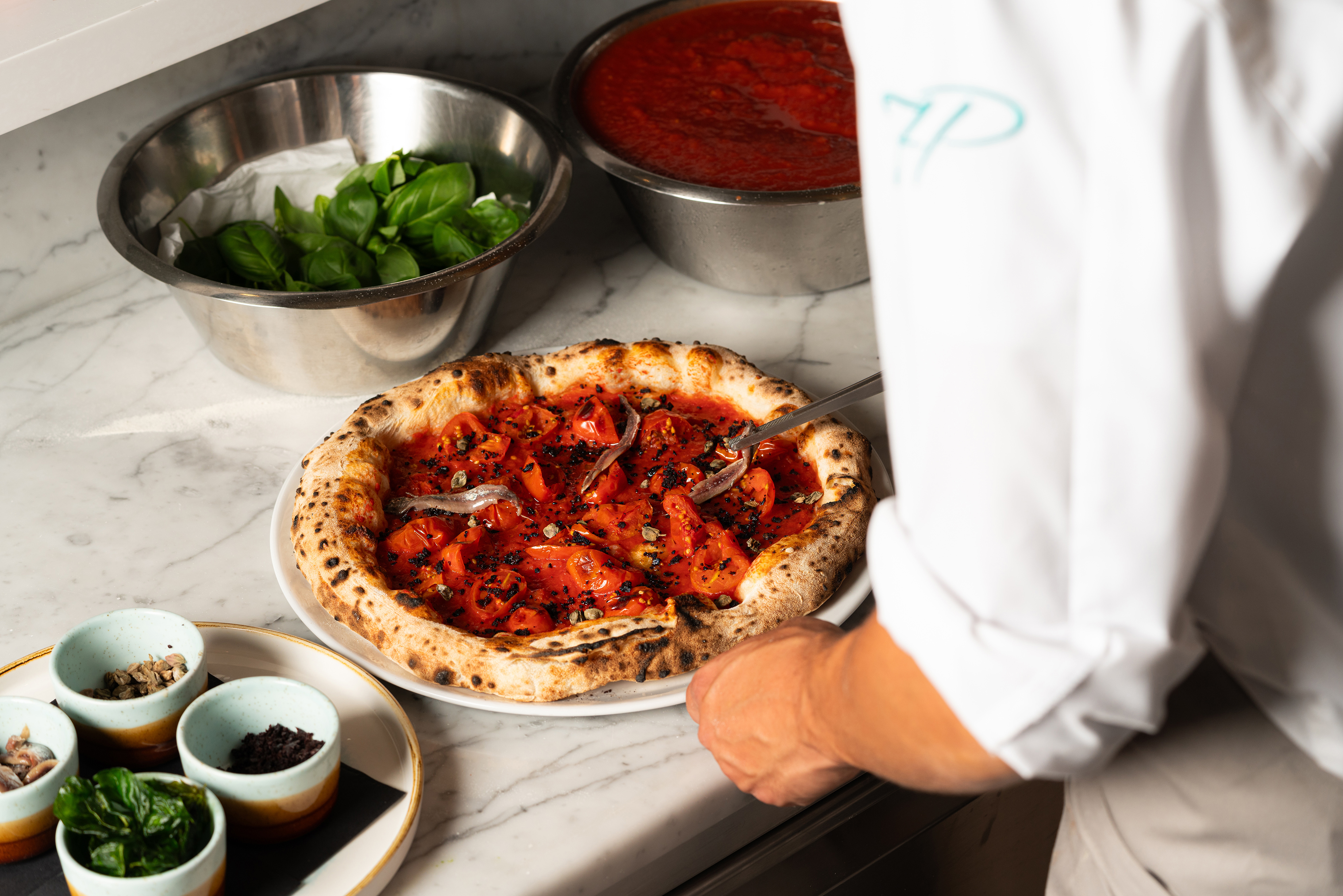 A chef making a stonebaked pizza surrounded by ingredients on top of a marble table