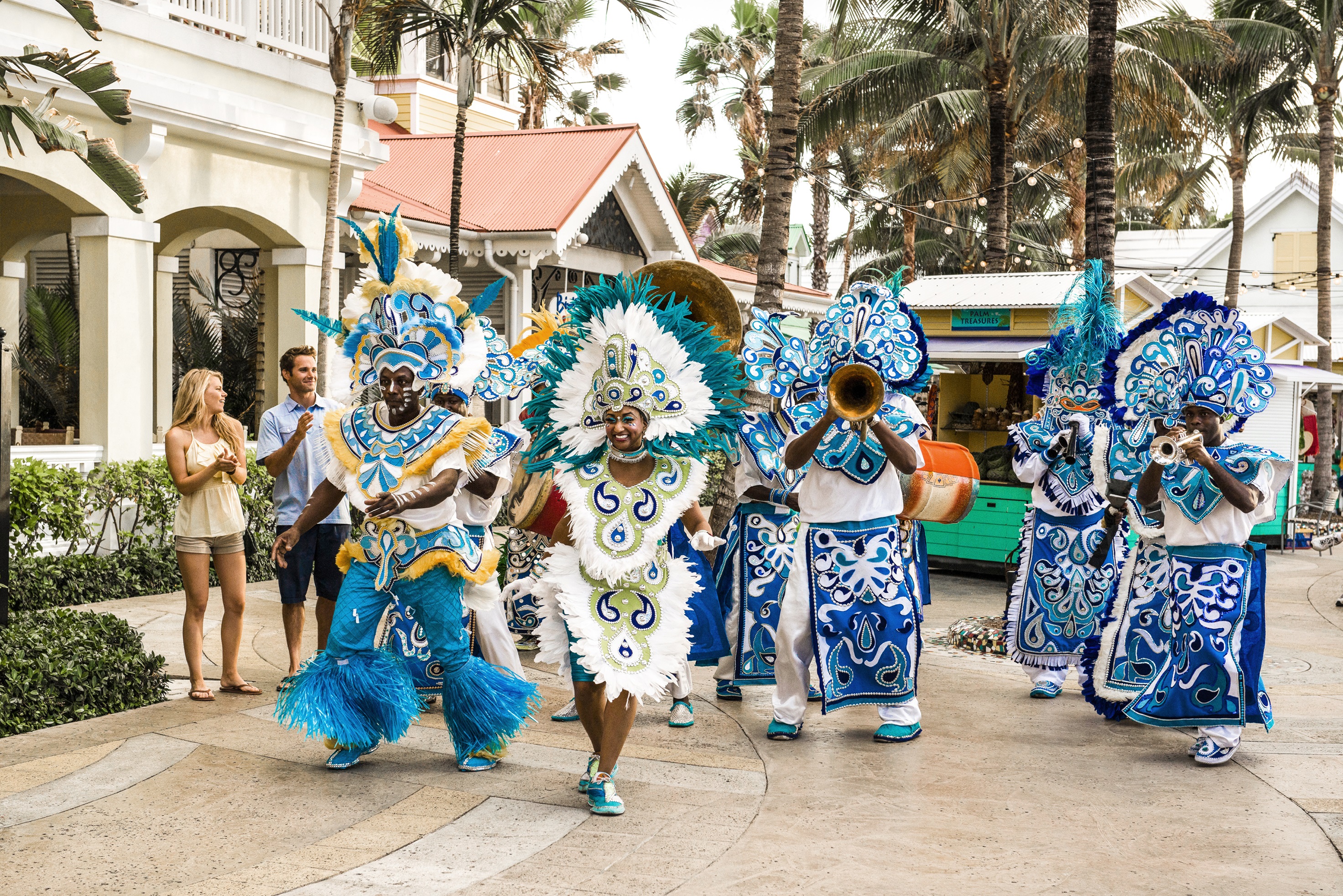 A colourful street parade in the Caribbean features dancers in bright blue and white costumes, playing brass instruments and smiling, while families watch and clap along in delight