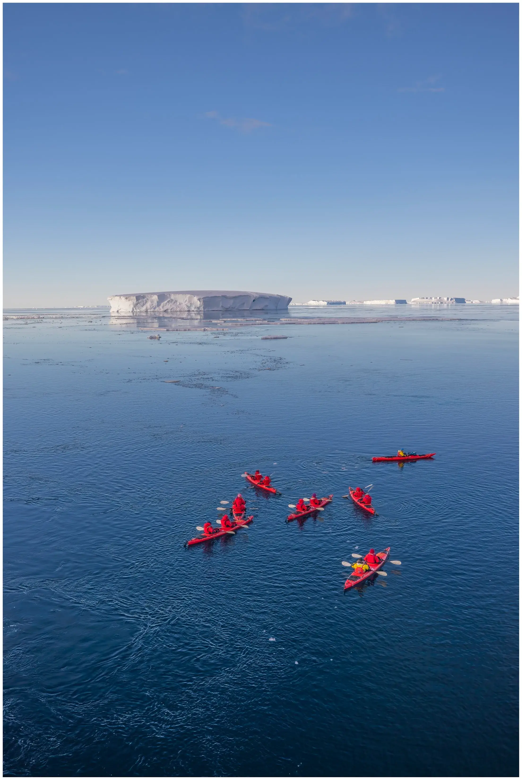 Ponant guests kayaking in icy Antarctic waters with a towering iceberg in the background under a clear sky.
