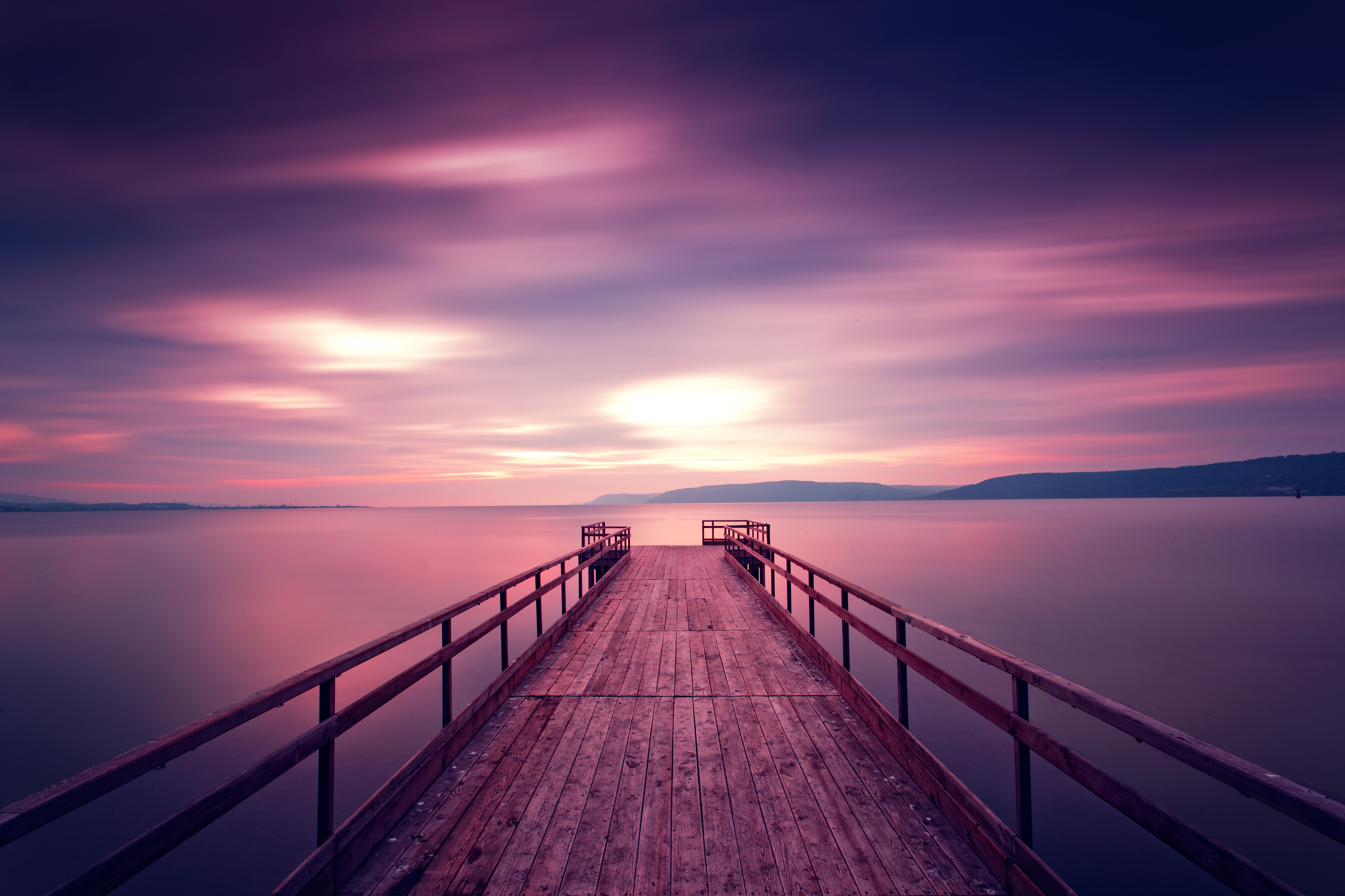 An empty dock stretches out into a pink and purple sunset, the colours of the sky reflecting on the water below