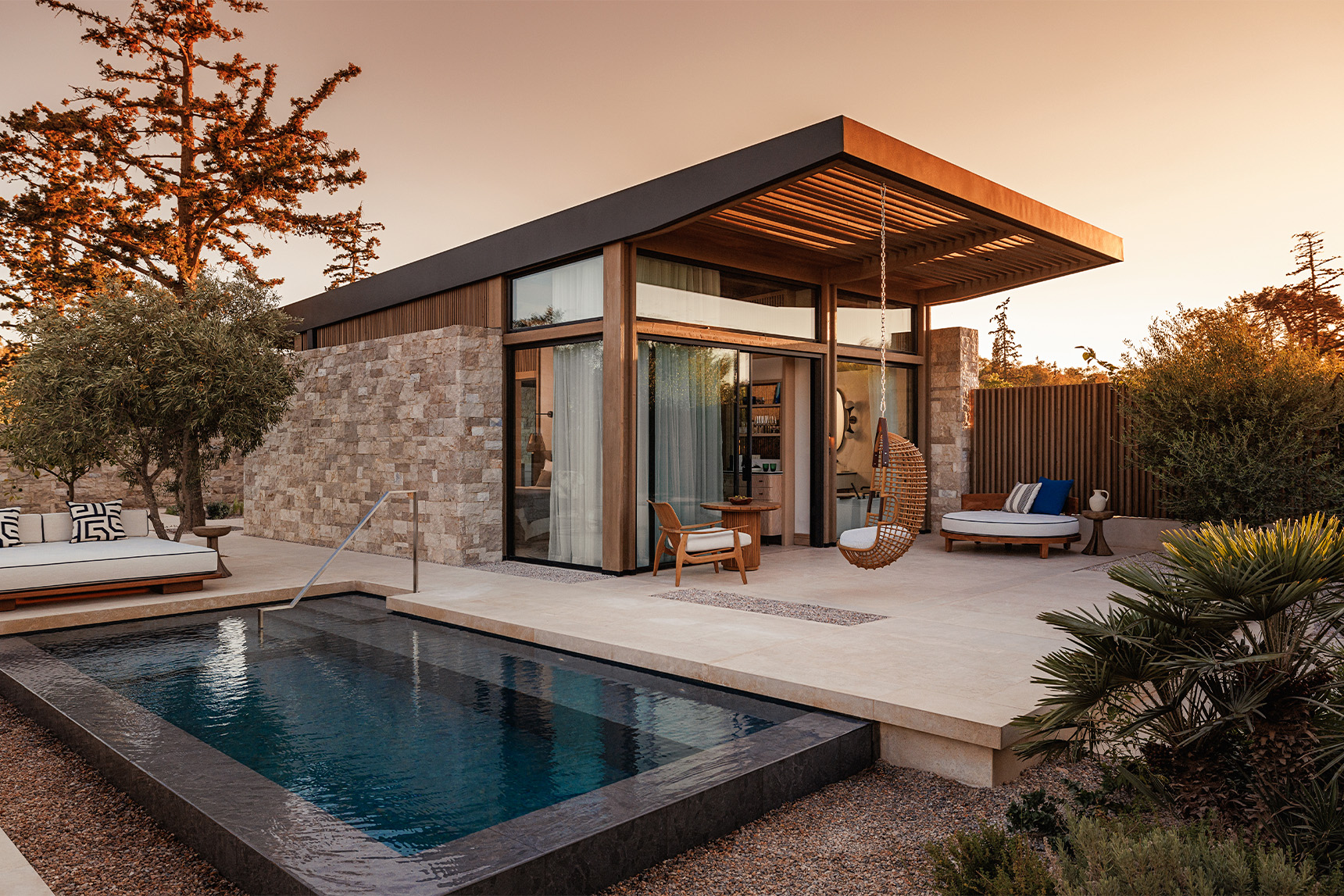 A chic stone bungalow with a small pool and patio with plush chairs is surrounded by green plants