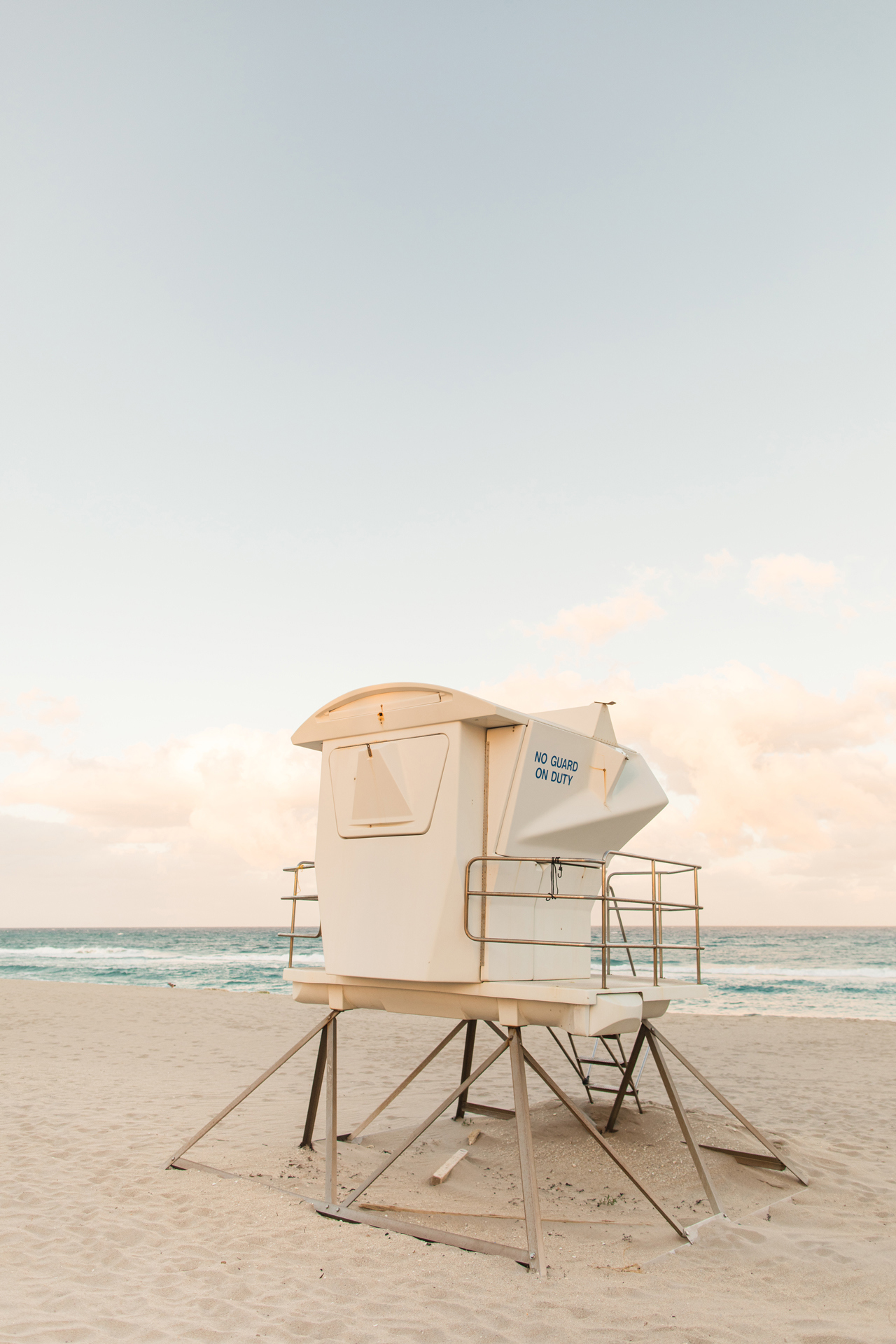 A white lifeguard stand on a sandy beach overlooking a pastel sunset and teal beach waves