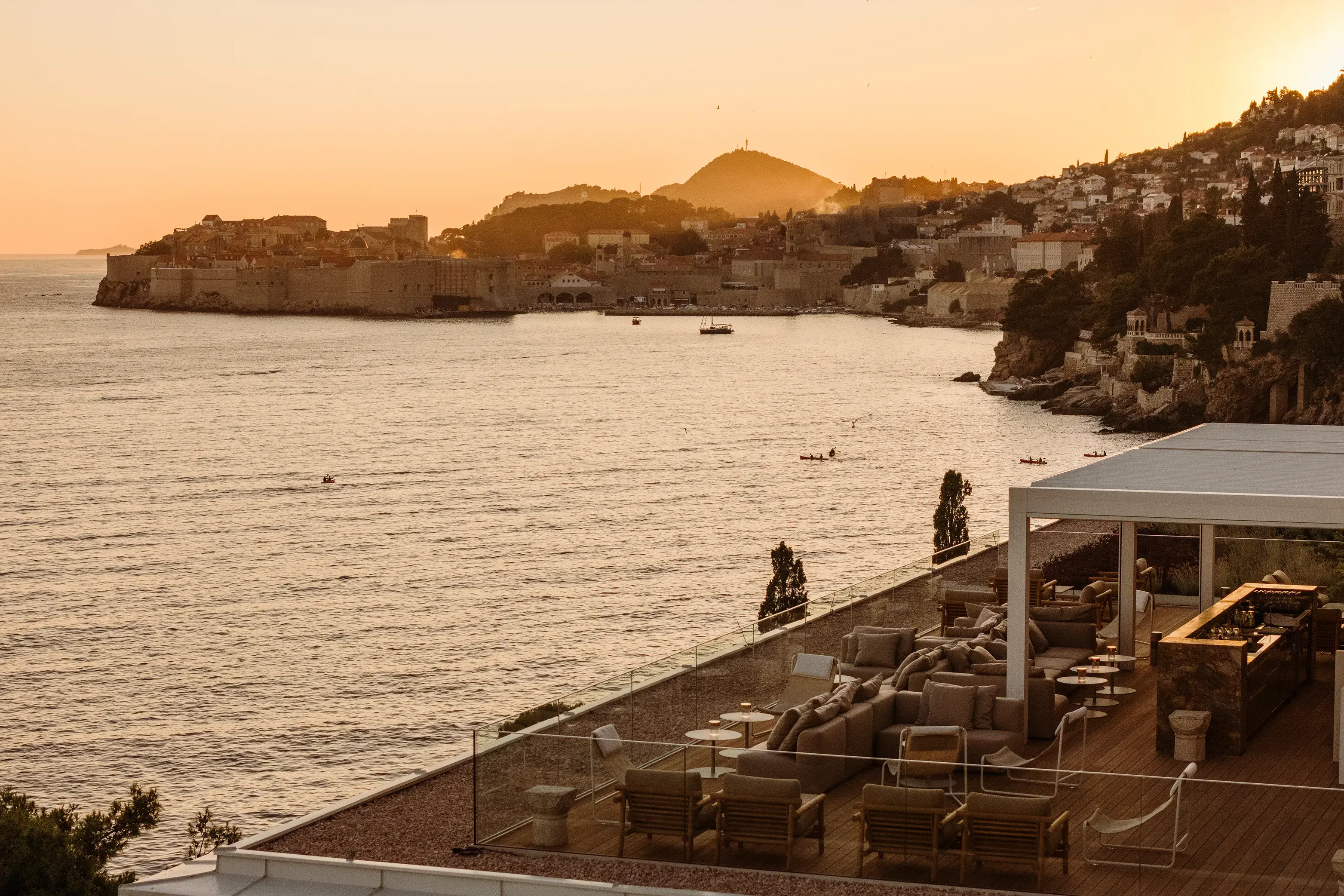 View of the rooftop Galanto Bar at Villa Dubrovnik in Croatia featuring a bar area with tables and chairs on a large terrace over the water with a view of the Old Town behind at sunset
