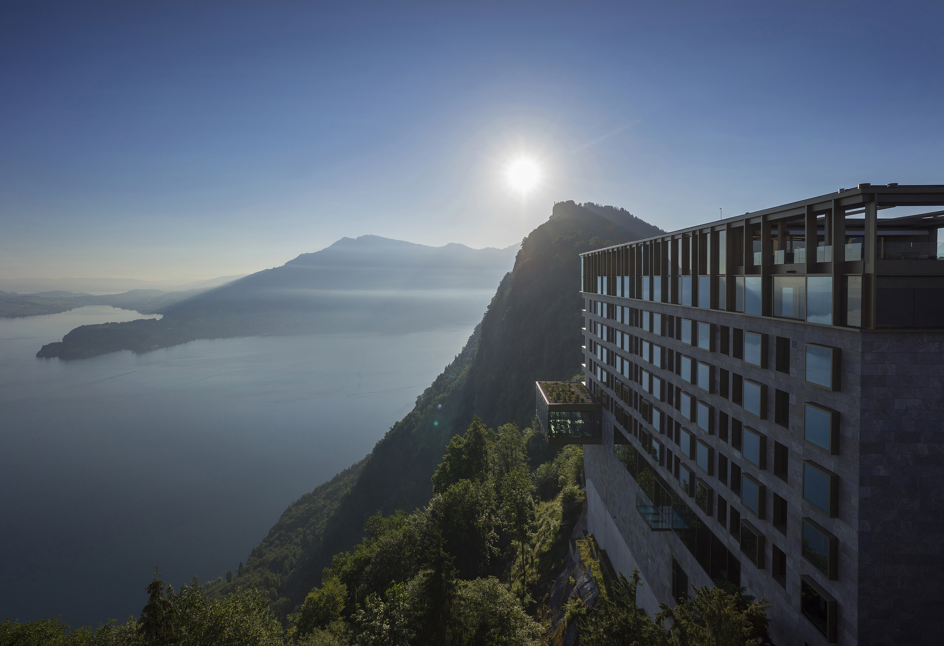 The exterior of Bürgenstock Hotel overlooking a sweeping hill and lake landscape with the sun shining in the background