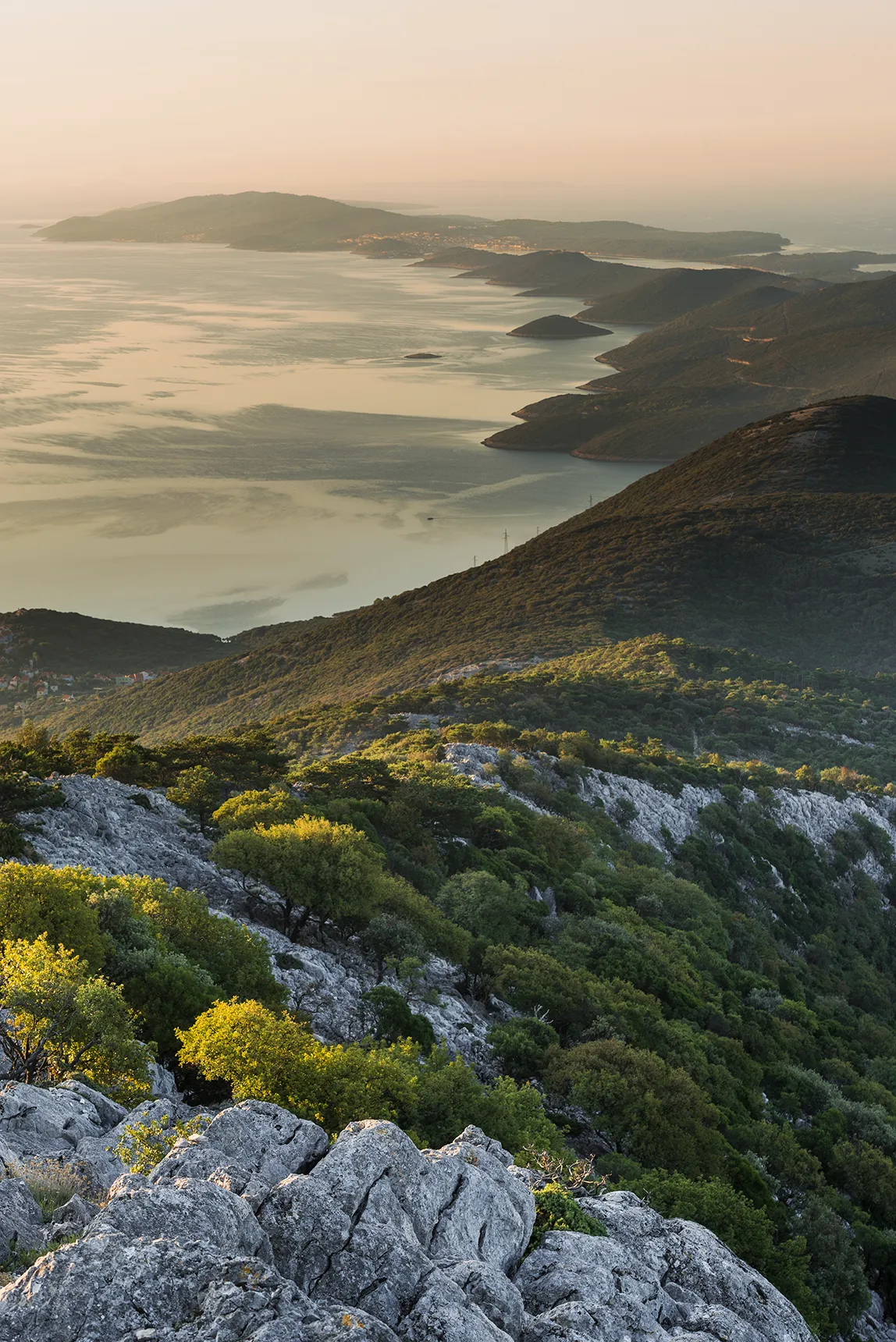 Mountainous coastline with forested hills and rocky slopes overlooking calm, island-dotted waters in soft evening light.