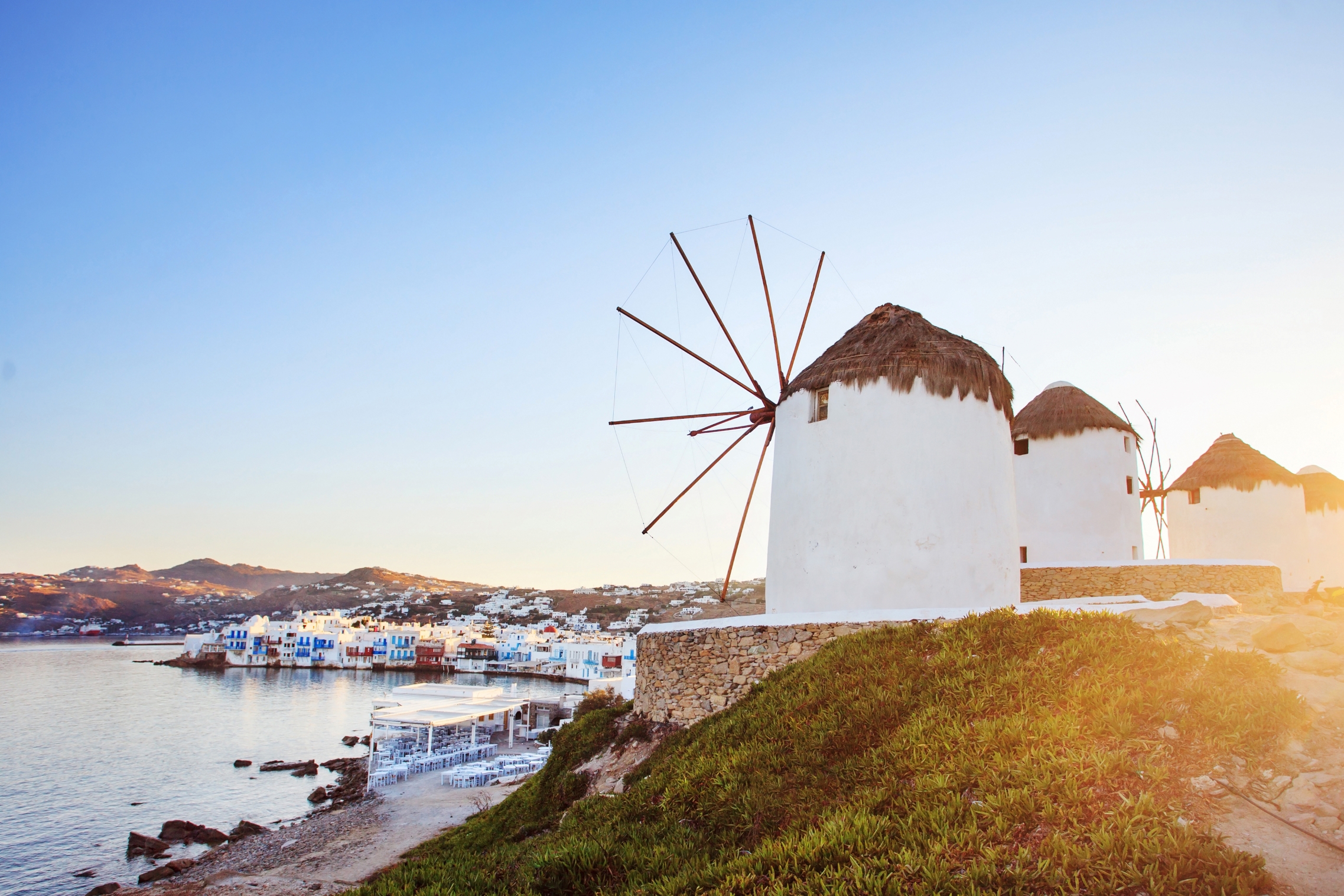 White-washed windmills overlook the colourful coast of Mykonos as mountains are seen in the distance