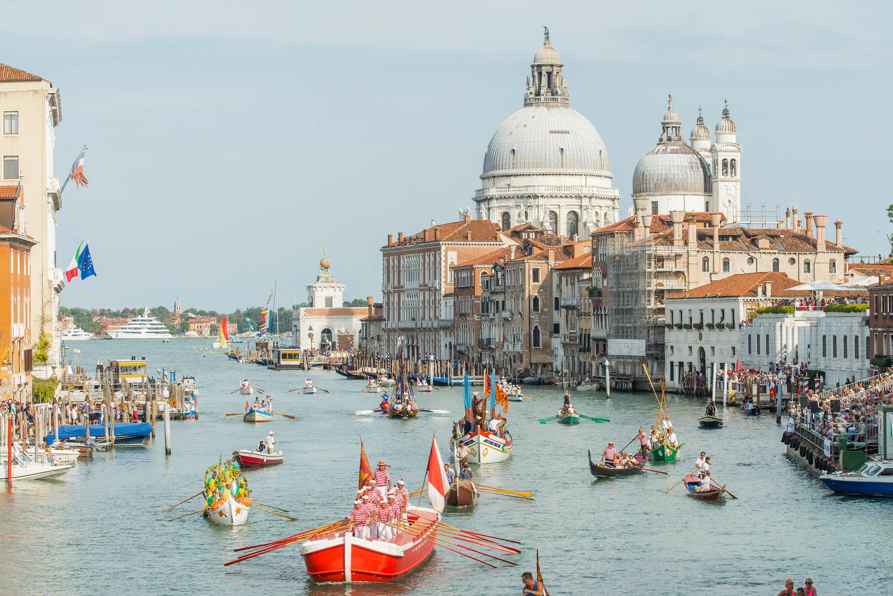Colourful gondolas and traditional boats parading along Venice Grand Canal with historic buildings and Santa Maria della Salute basilica in the background.