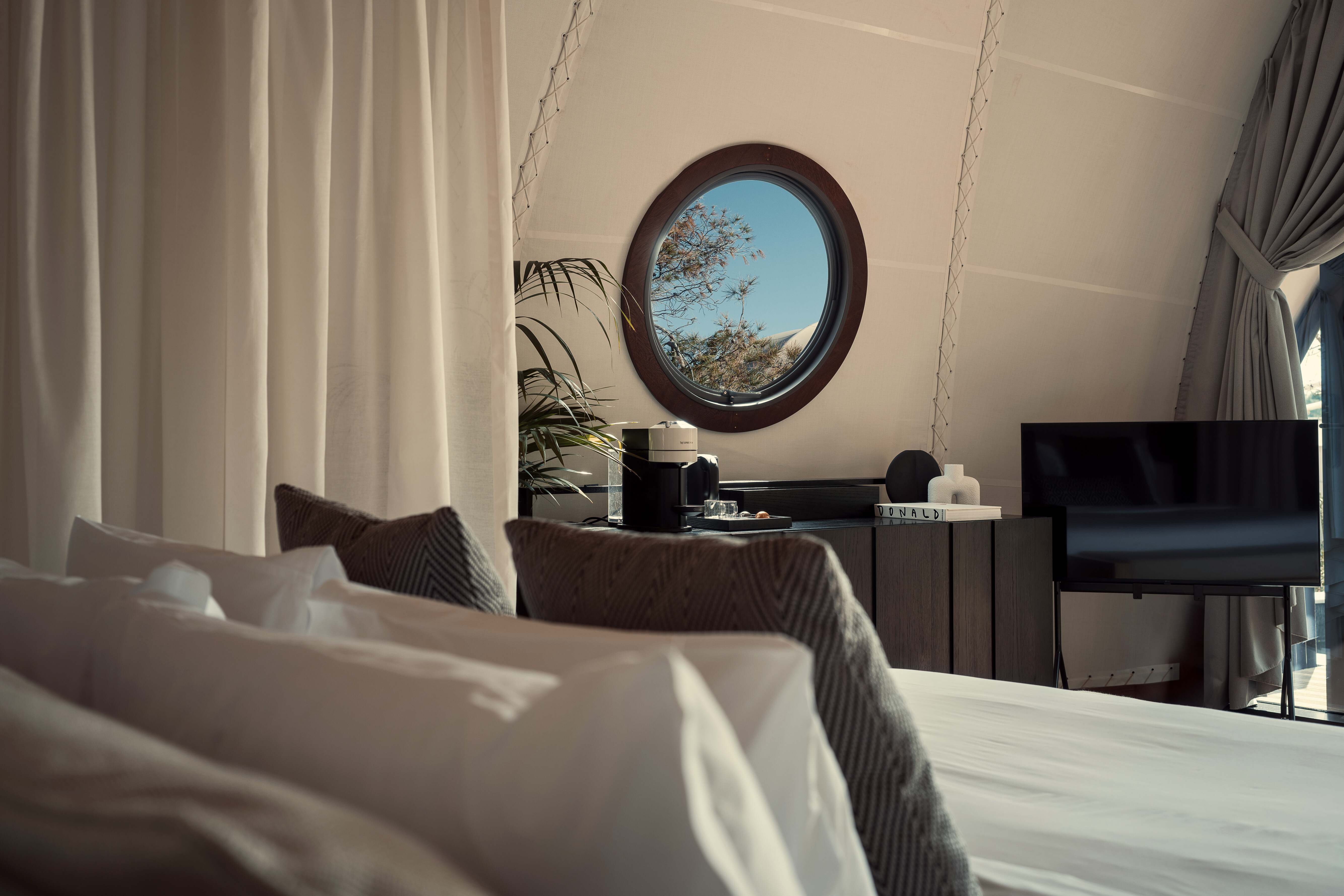 Bedroom sideboard with decorative items on top below a circular window beside a TV with plush bed cushions in soft focus
