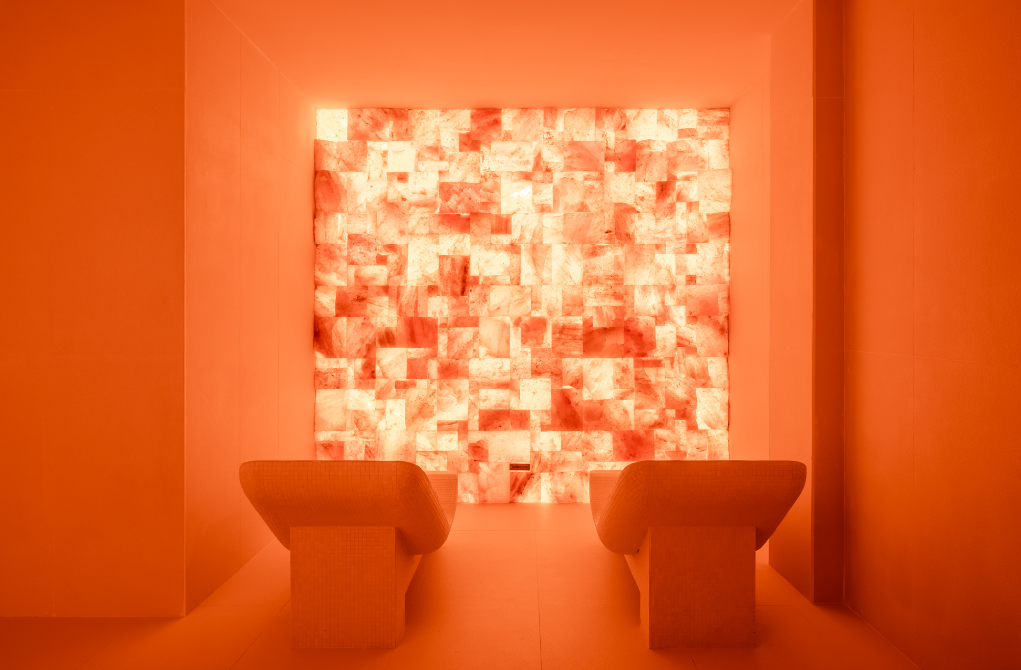 A warmly lit room with two beige chairs facing a large, illuminated wall of textured pink and white blocks.