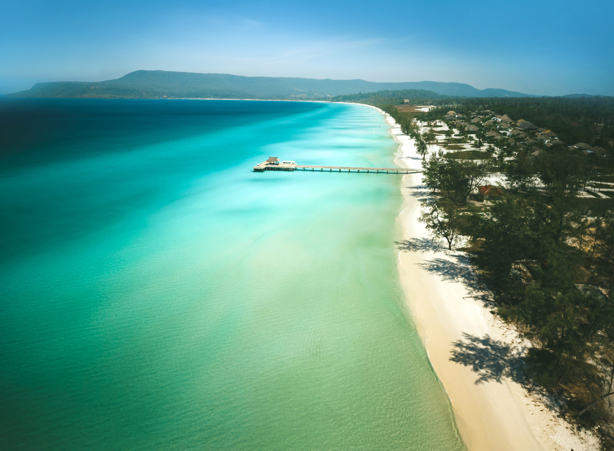 Aerial view of a turquoise sea with a long pier, sandy beach, and coastal buildings under a clear sky.