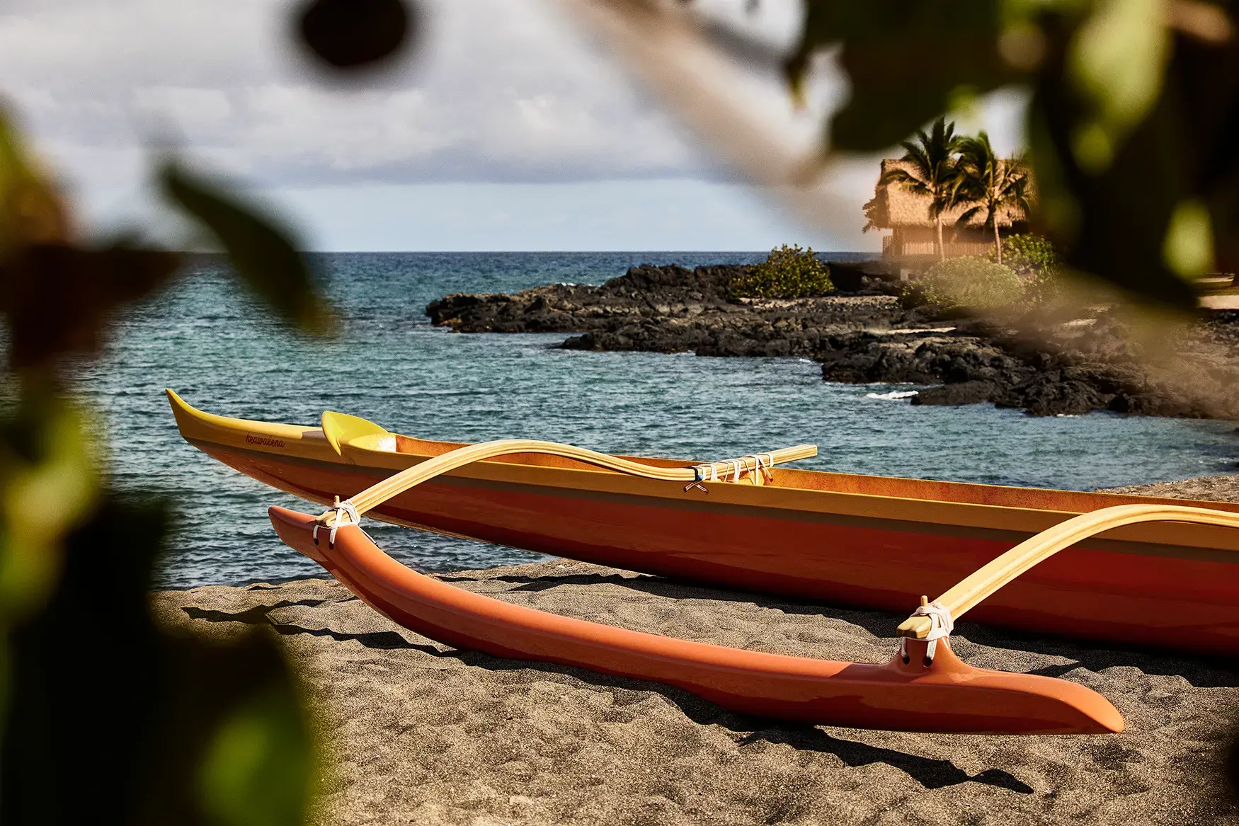 An traditional orange canoe sitting on the beach beside the ocean with blurred foliage in the foreground