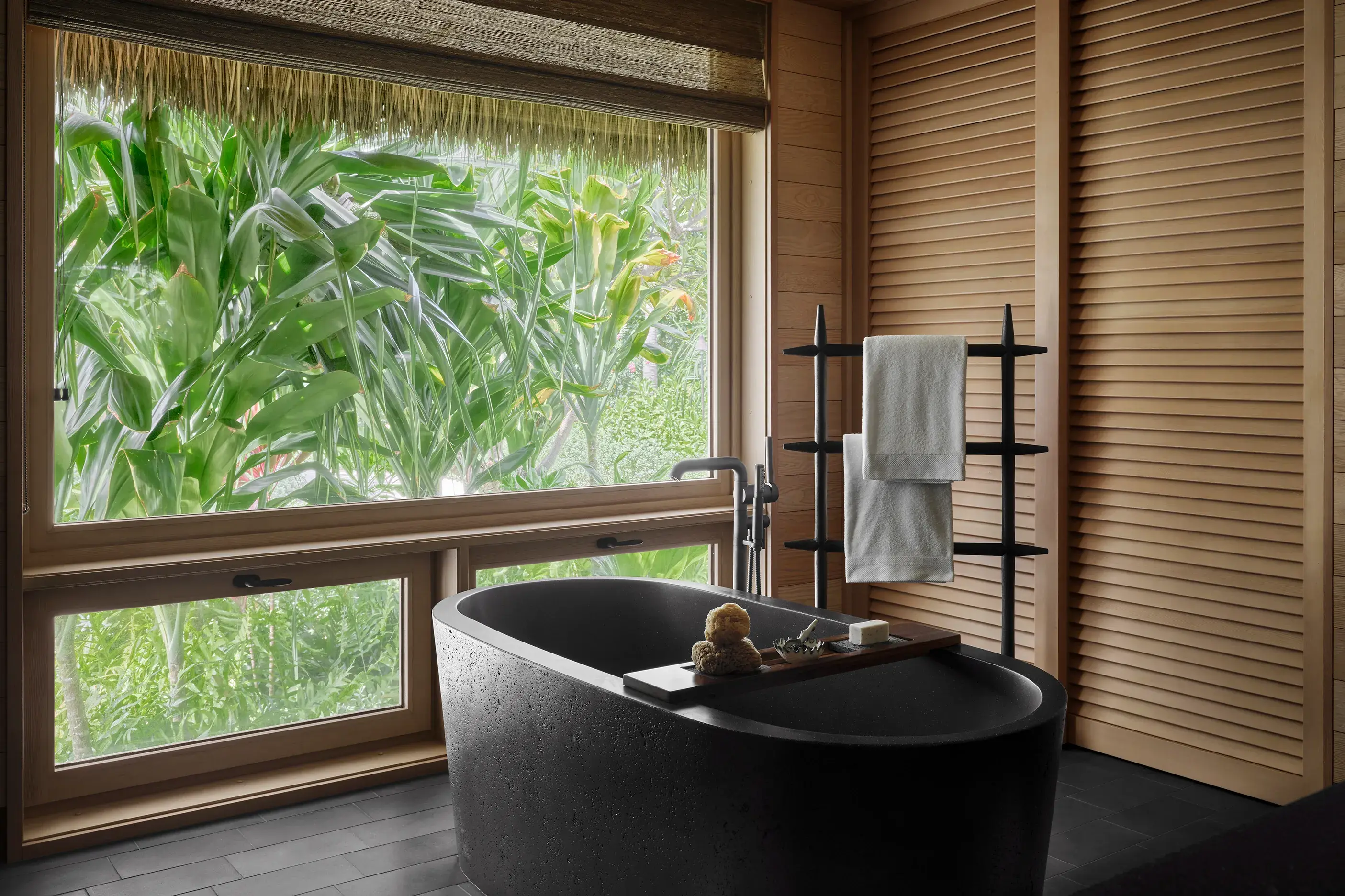 The sleek bathroom of a Kauhale Beachfront bathroom featuring a large black bathtub and wood paneled walls looking out large windows onto dense foliage
