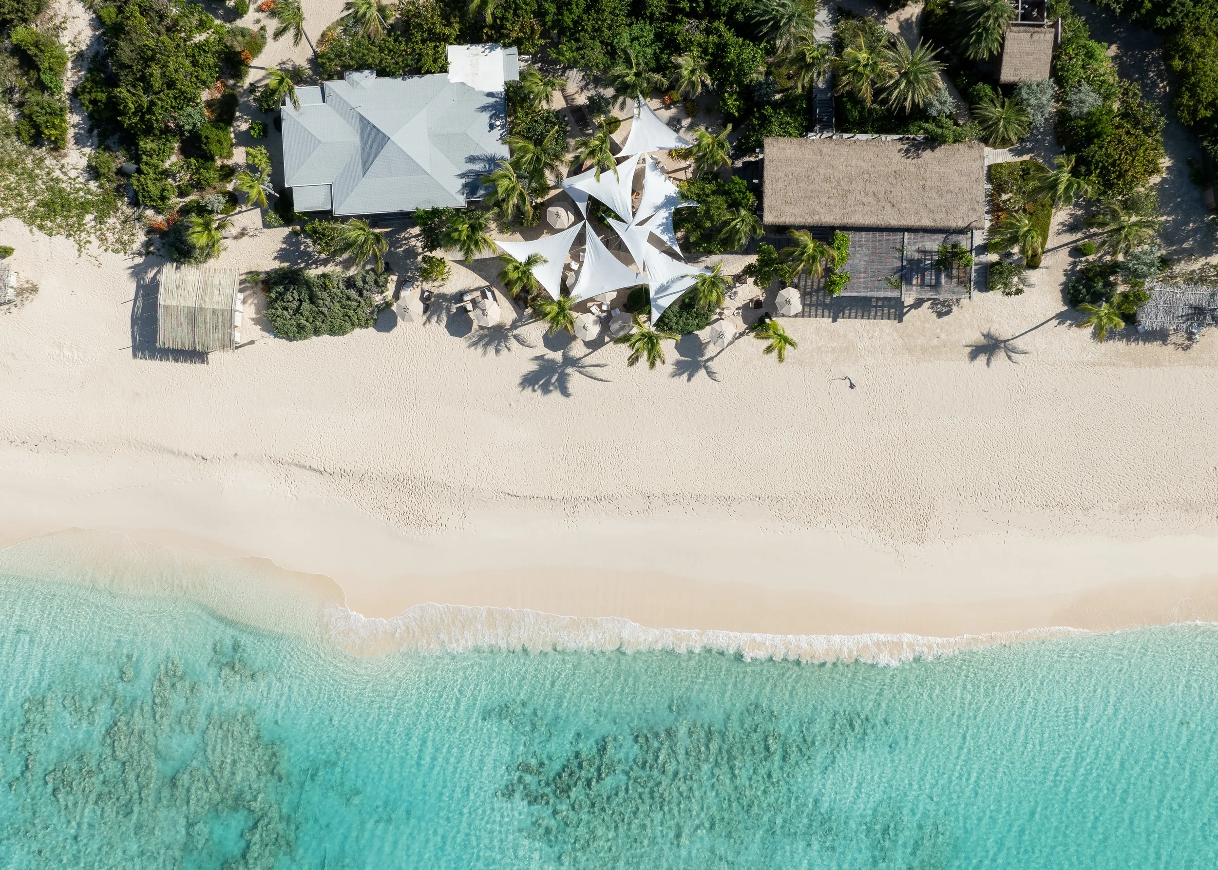 Aerial view of a tropical beachfront Nobu Barbuda resort with white sand, turquoise water, palm trees, and shaded lounge areas.