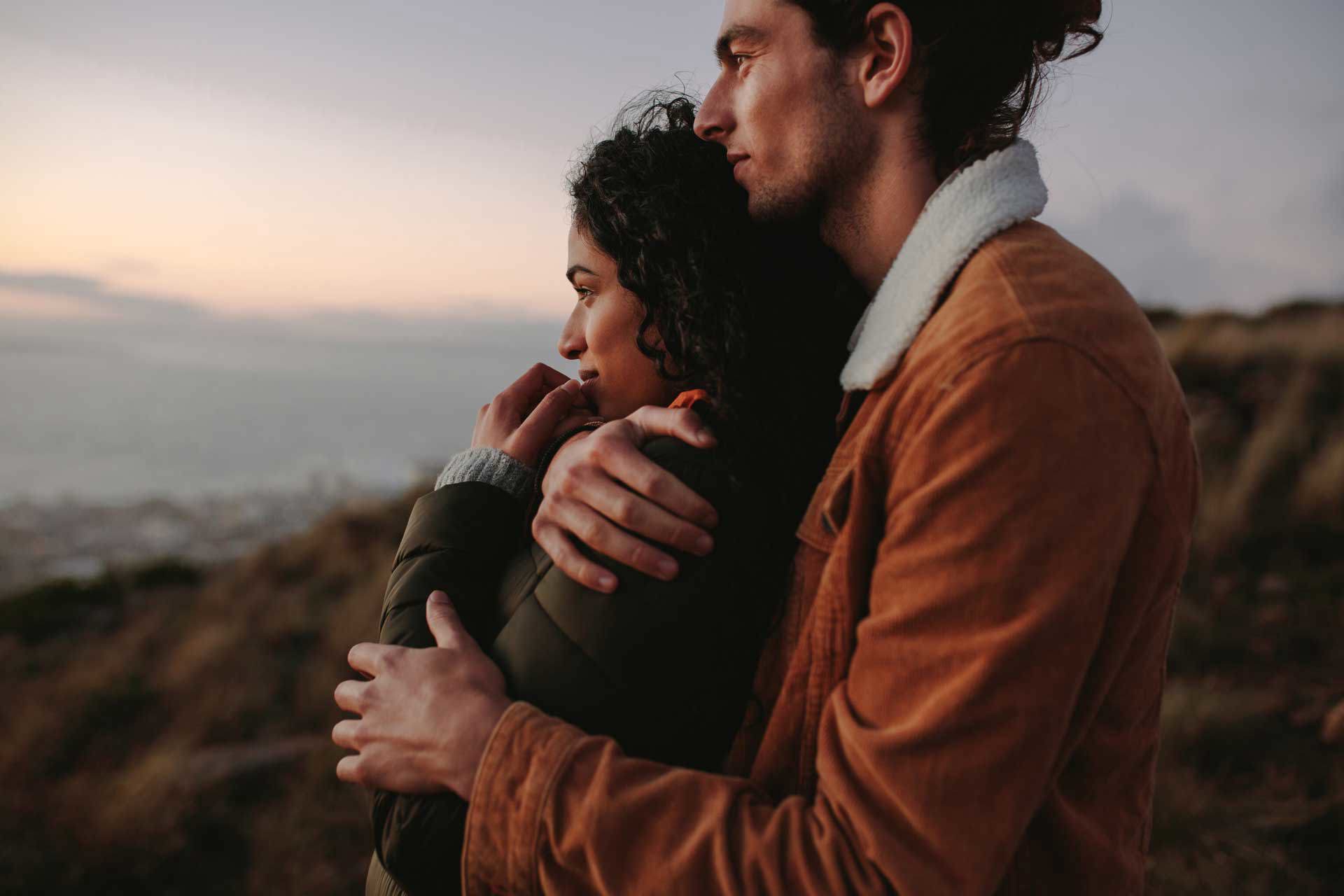 Two people standing close together on a coastal cliff, wrapped in jackets and looking out over the sea at sunset.