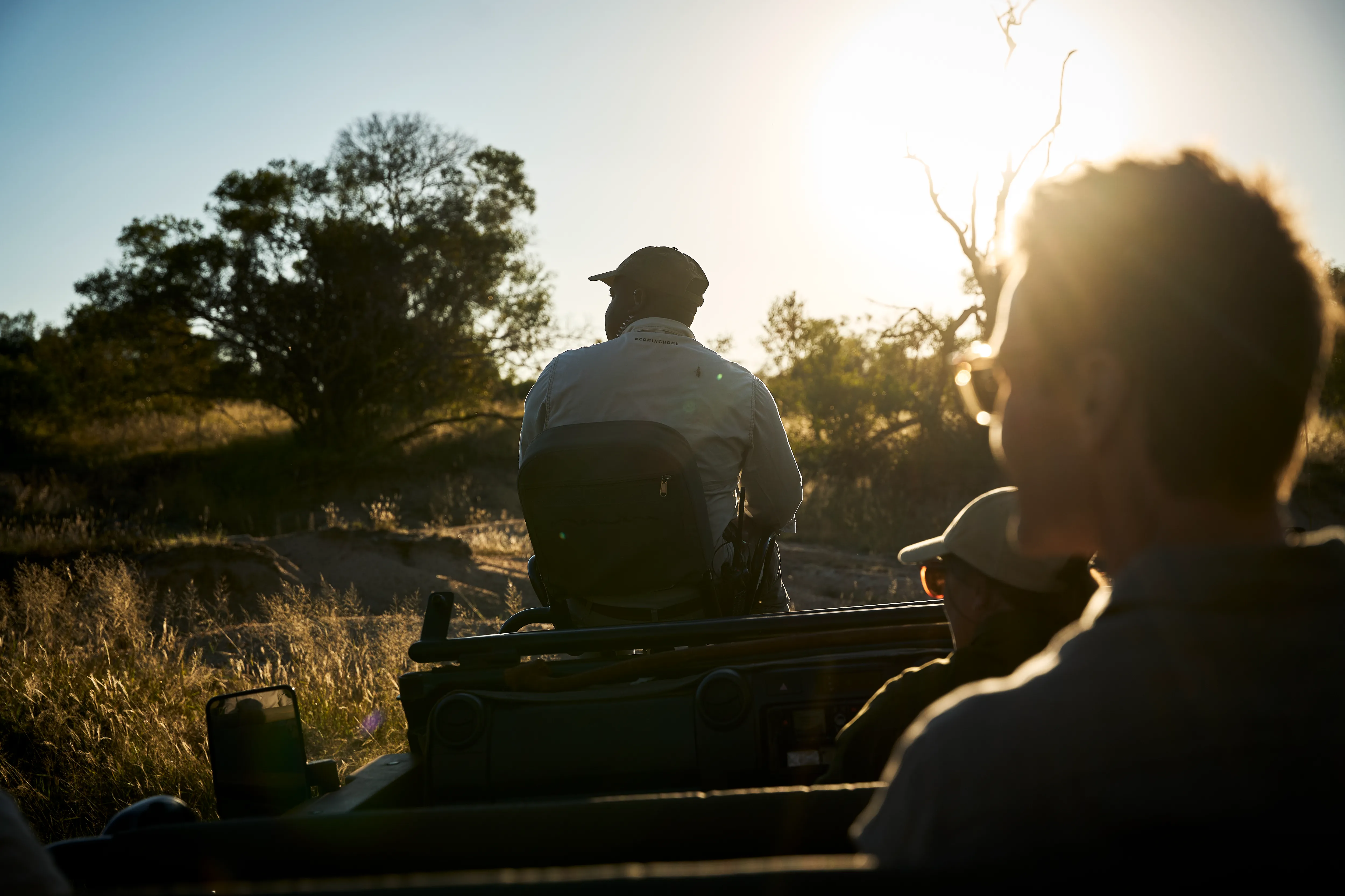 Three people aboard a safari jeep in the South African bush with the sun casting a flare over them