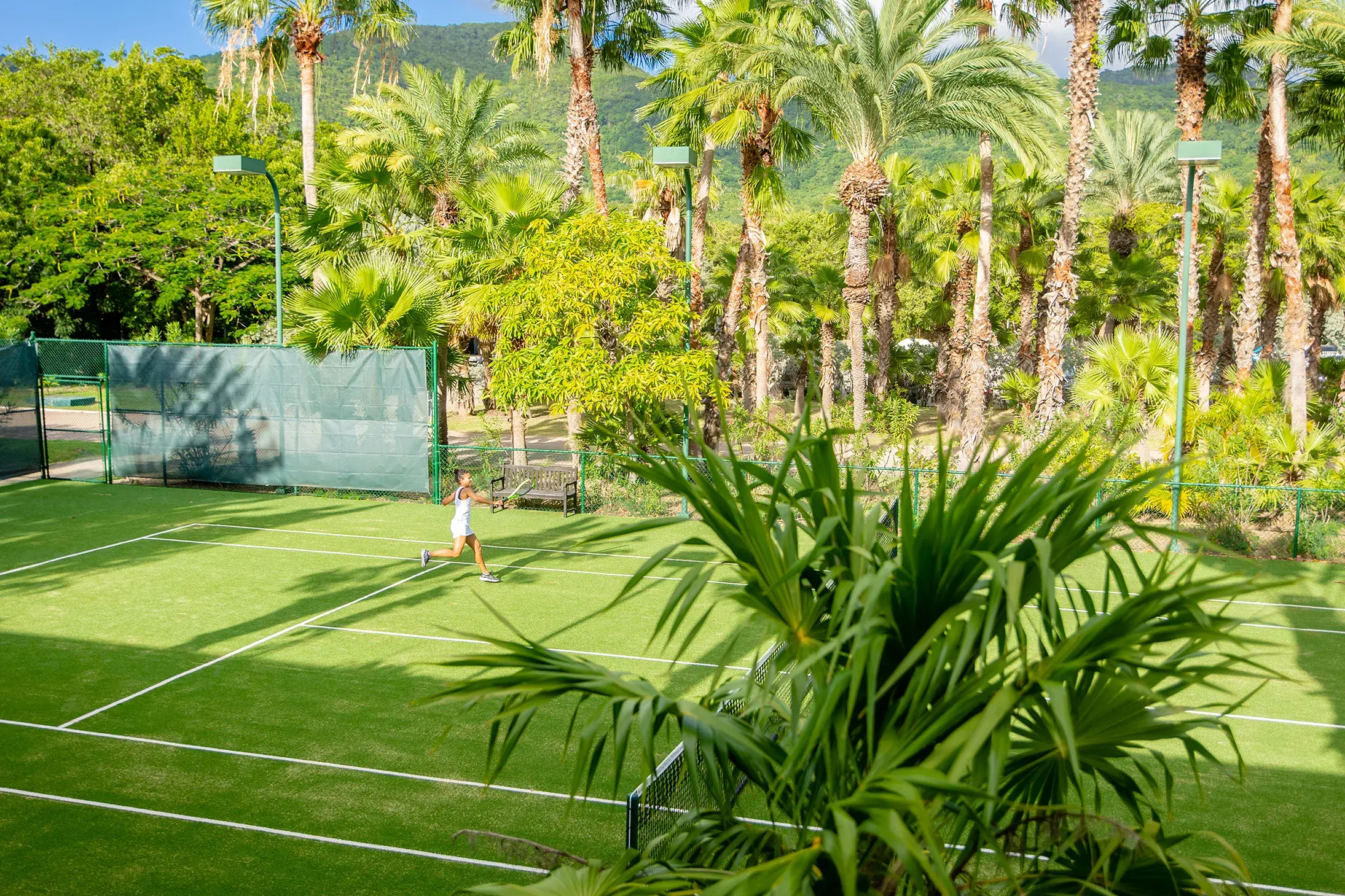 A guest playing tennis on the courts at Carlisle Bay surrounding by palm trees