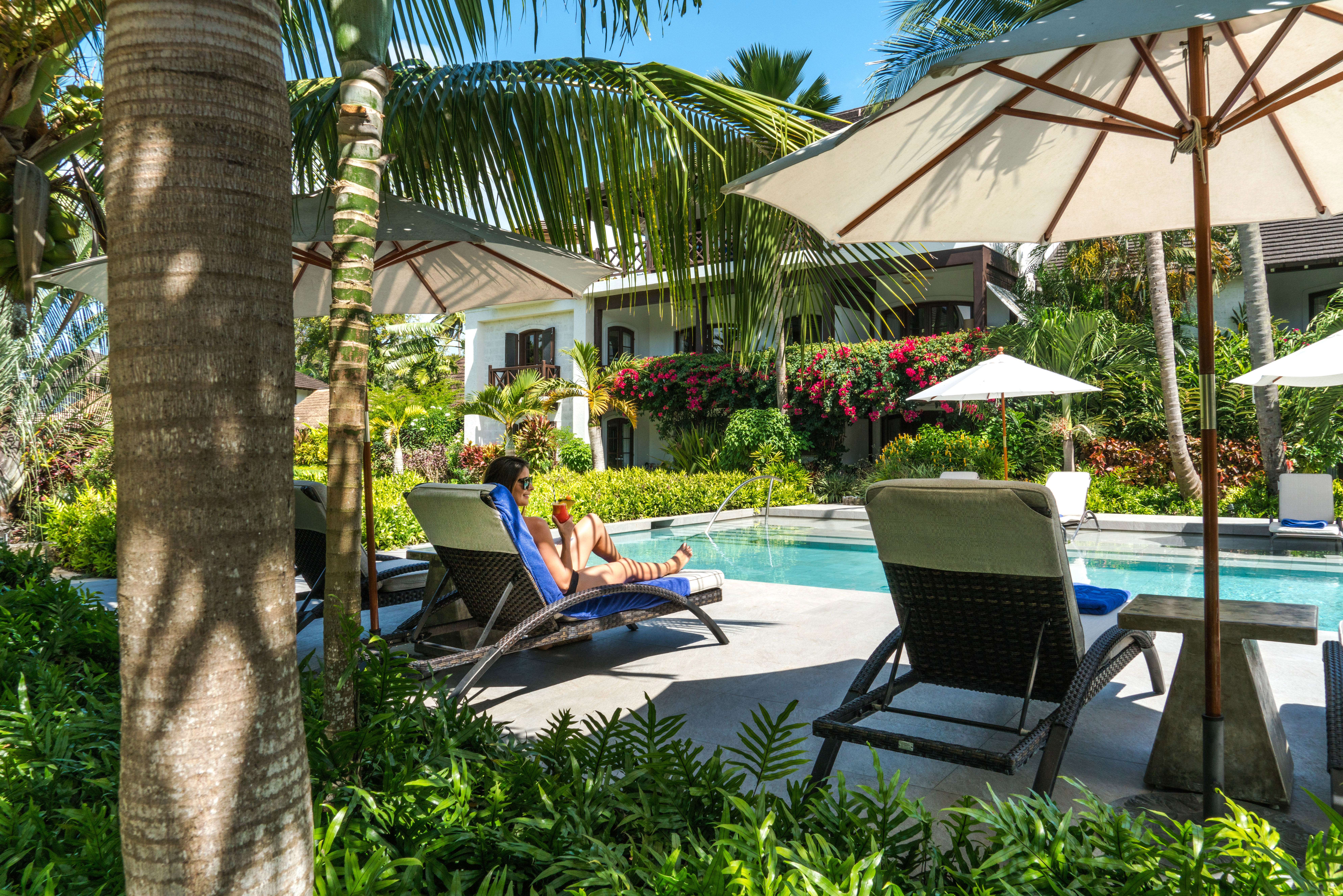 A woman with a drink lounging on a blue sun lounger beside a pool shaded by palms at The Sandpiper