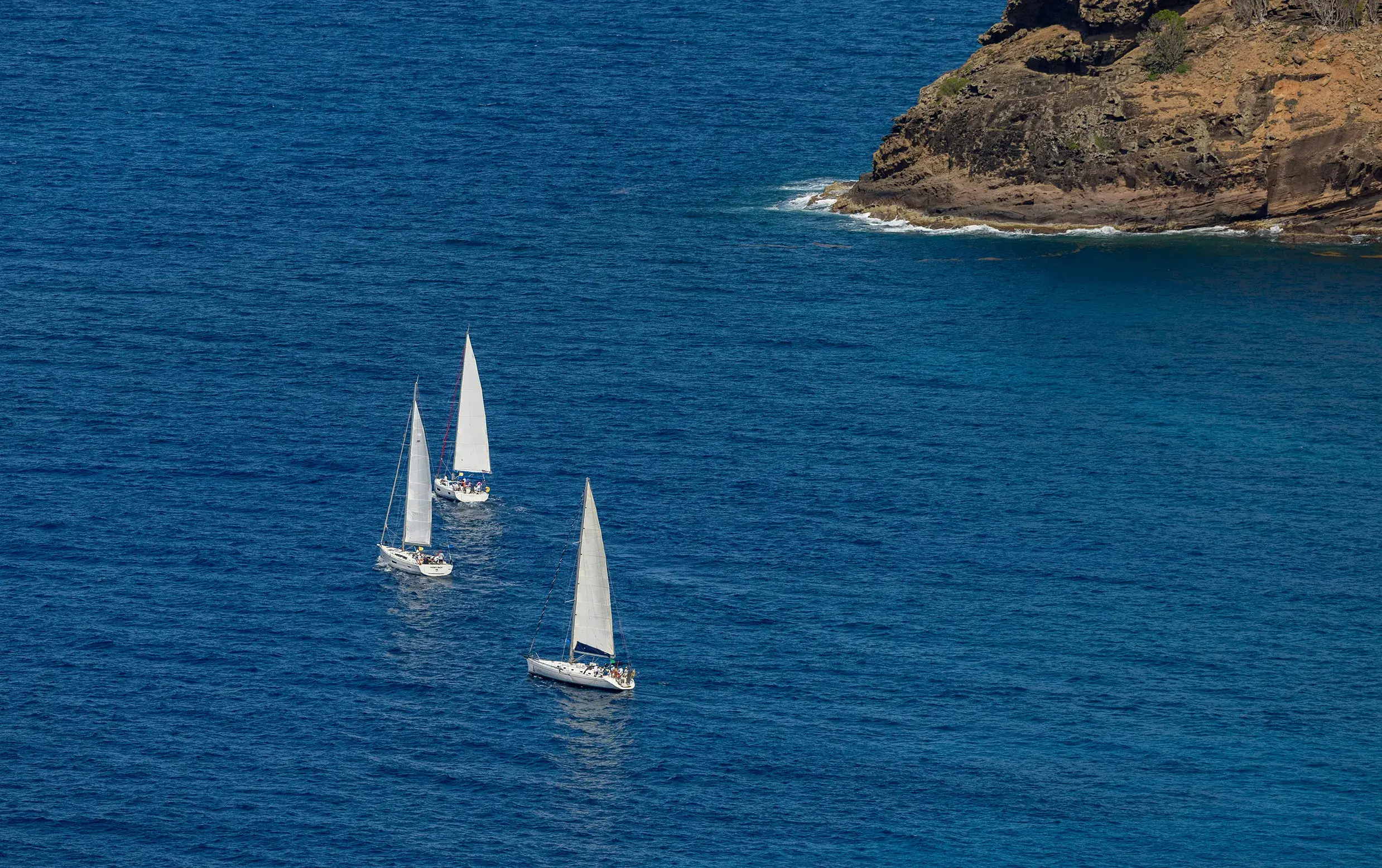 Three white sailboats gliding on deep blue ocean water near a rocky coastline.