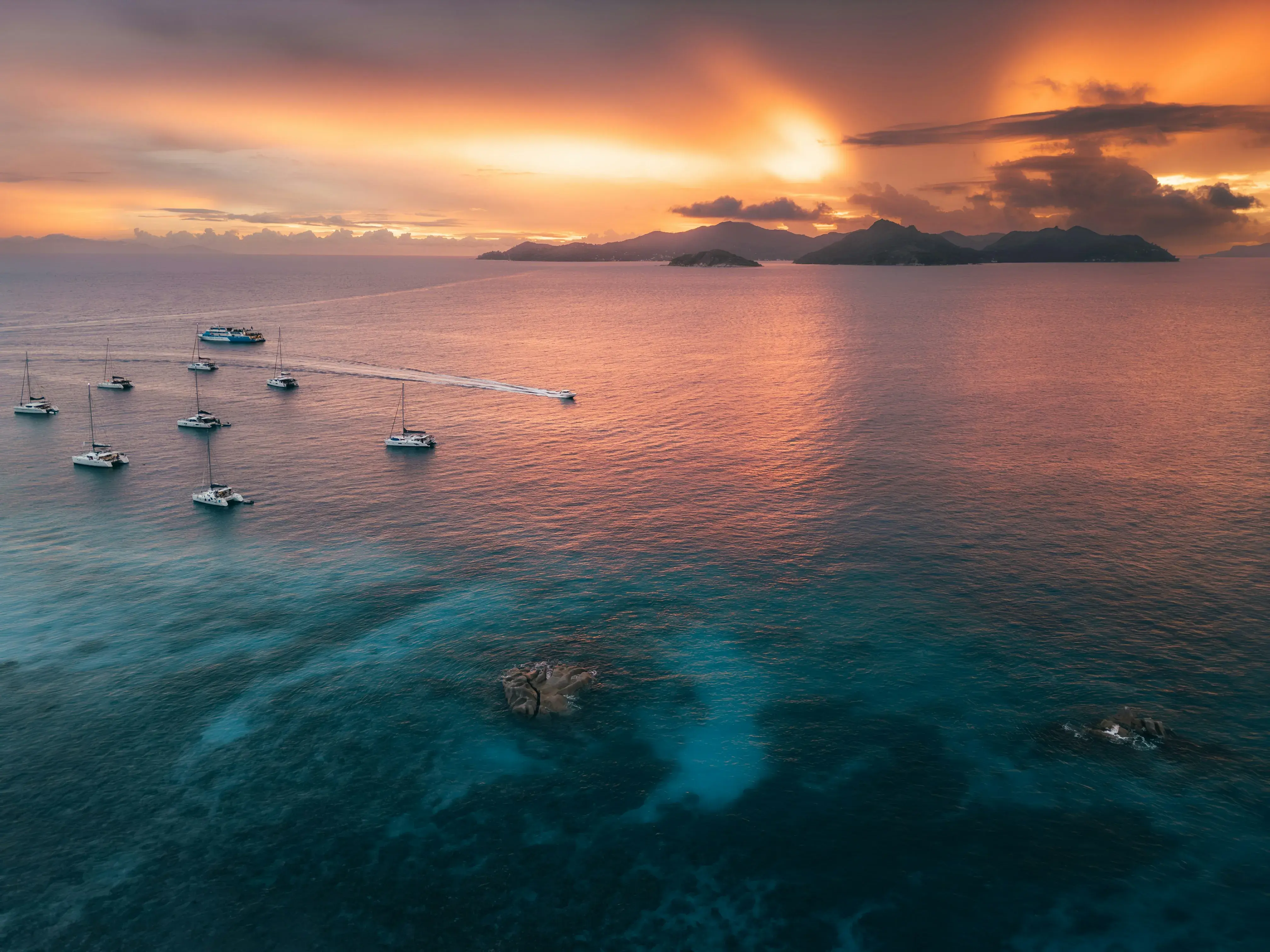 Sailboats anchored on clear blue water at sunset with vibrant orange sky and distant islands on the horizon in the Seychelles.