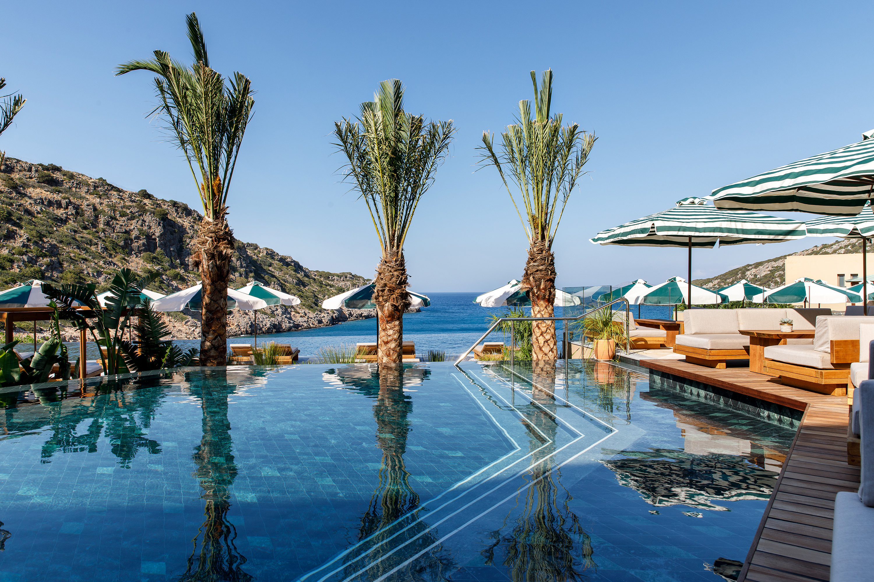 Calm swimming pool at Daios Cove fronted by palm trees with rows of striped sun umbrellas and loungers