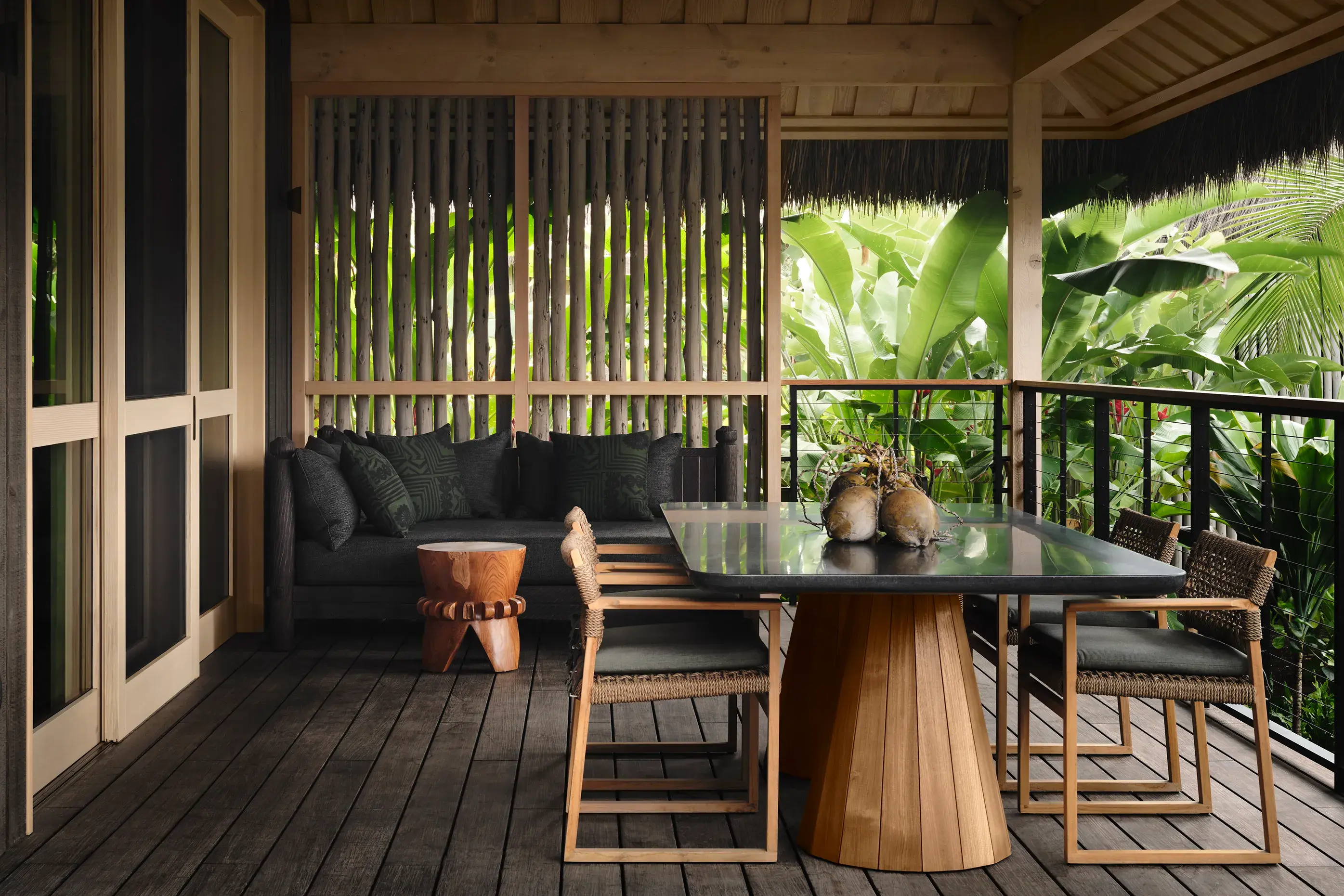 The terrace of a Kauhale Lagoon View featuring a wood and glass dining table and black sofa beneath a thatched roof surrounded by deep vegetation 