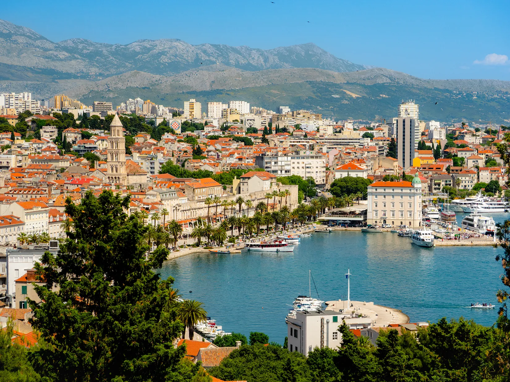 Harbor city with red-roofed buildings and mountains in the background.
