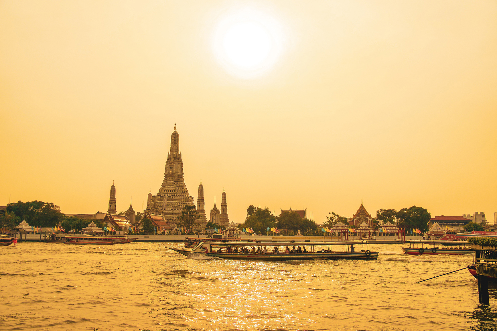 Wat Arun Ratchawararam Ratchawaramahawihan temple in Bangkok at sunset with boats on the river in foreground
