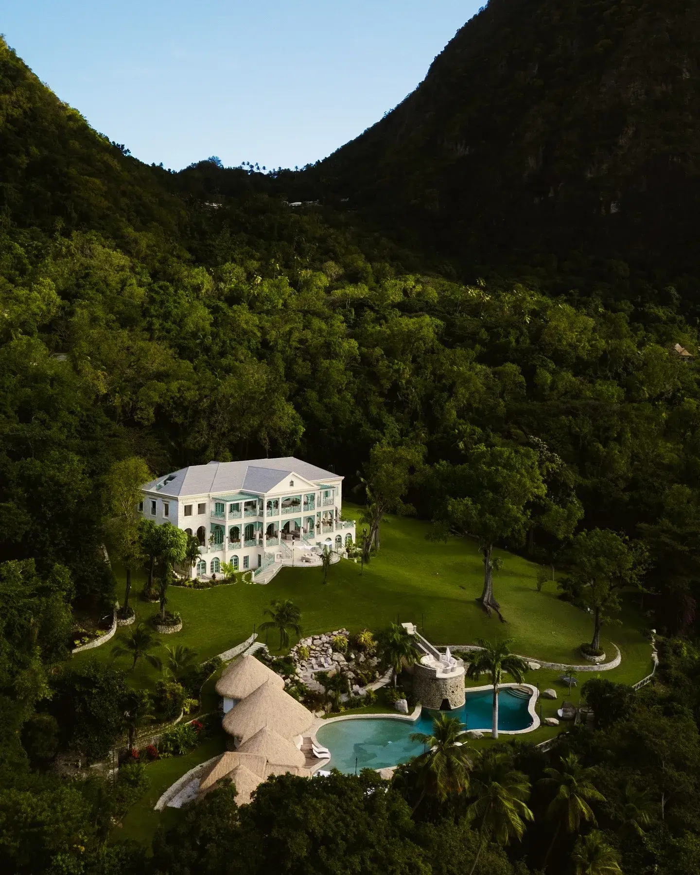 Aerial view of La Belle Helene villa in St Lucia, surrounded by lush green hills with a pool and thatched-roof structures in the foreground.