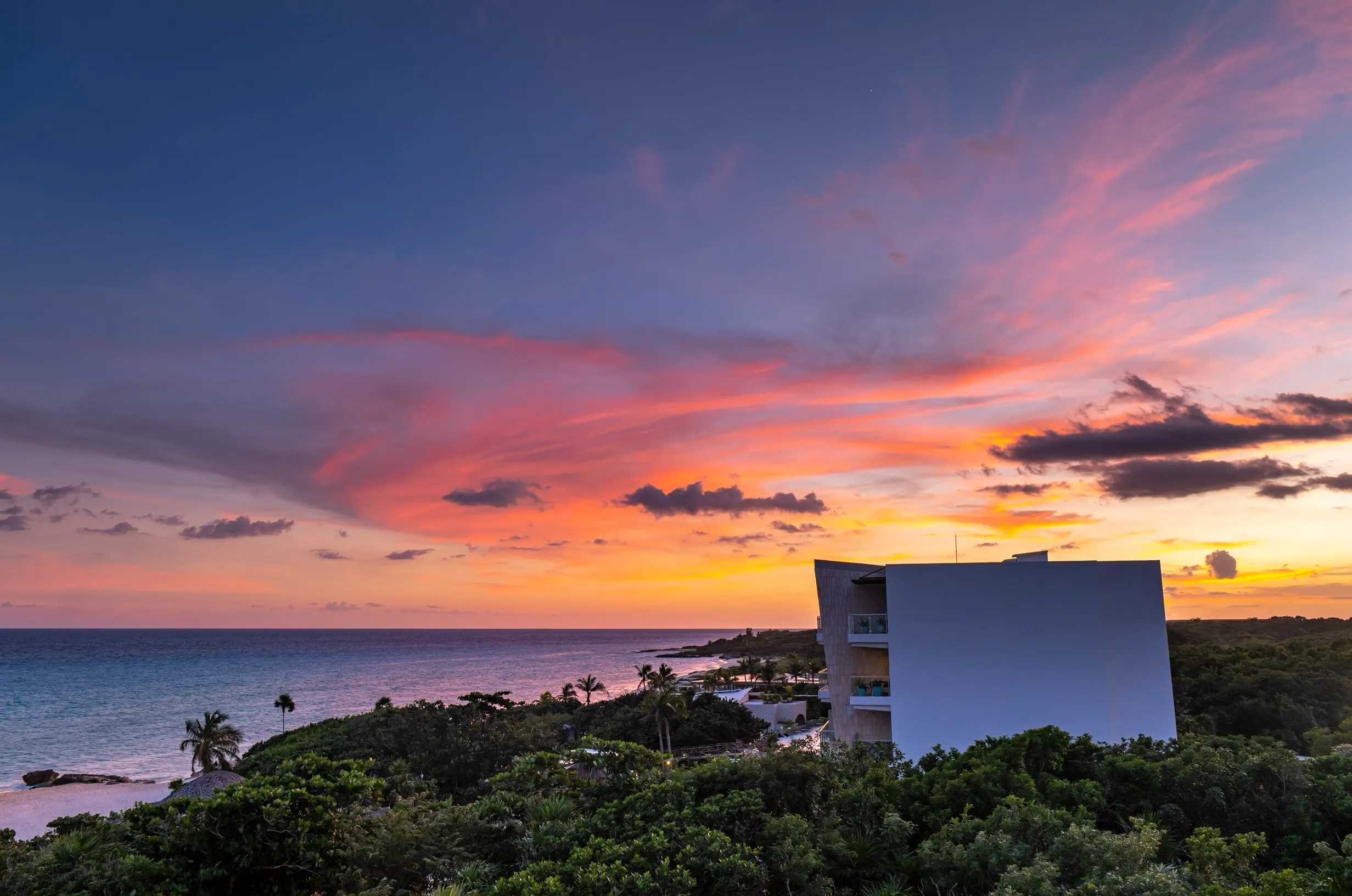 Colourful sunset over ocean with modern resort building on a tropical coastline.