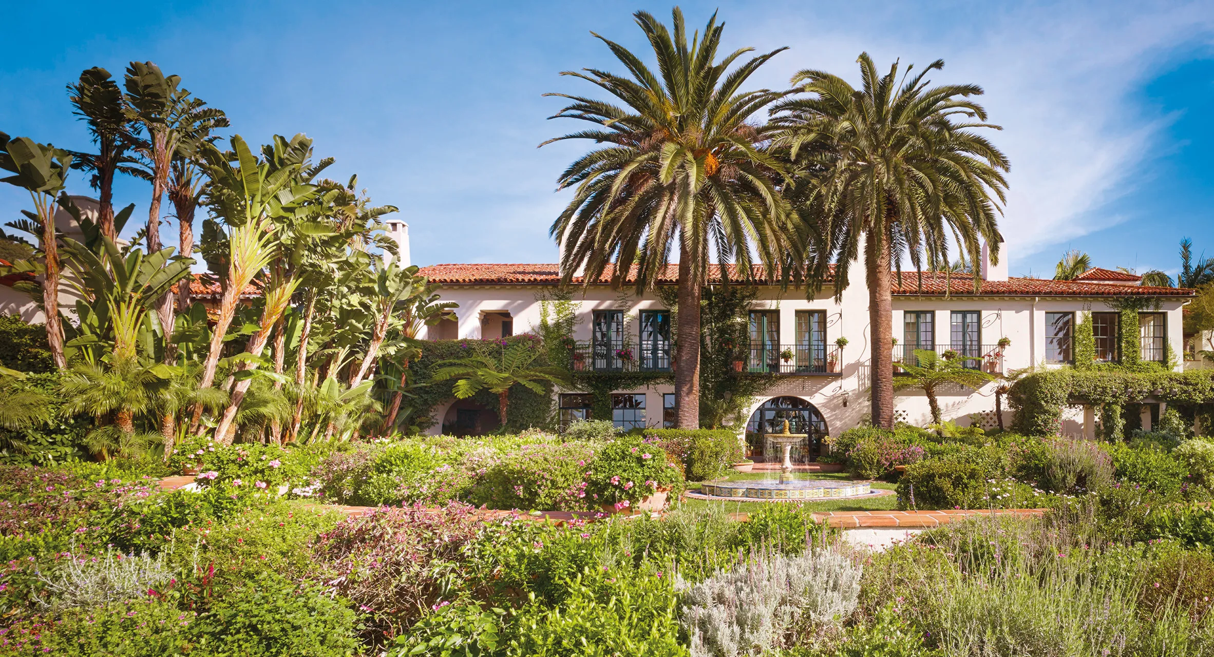 Luxury Spanish-style villa of Four Seasons Resort The Biltmore surrounded by lush tropical gardens, palm trees, and a central fountain under a clear blue sky.