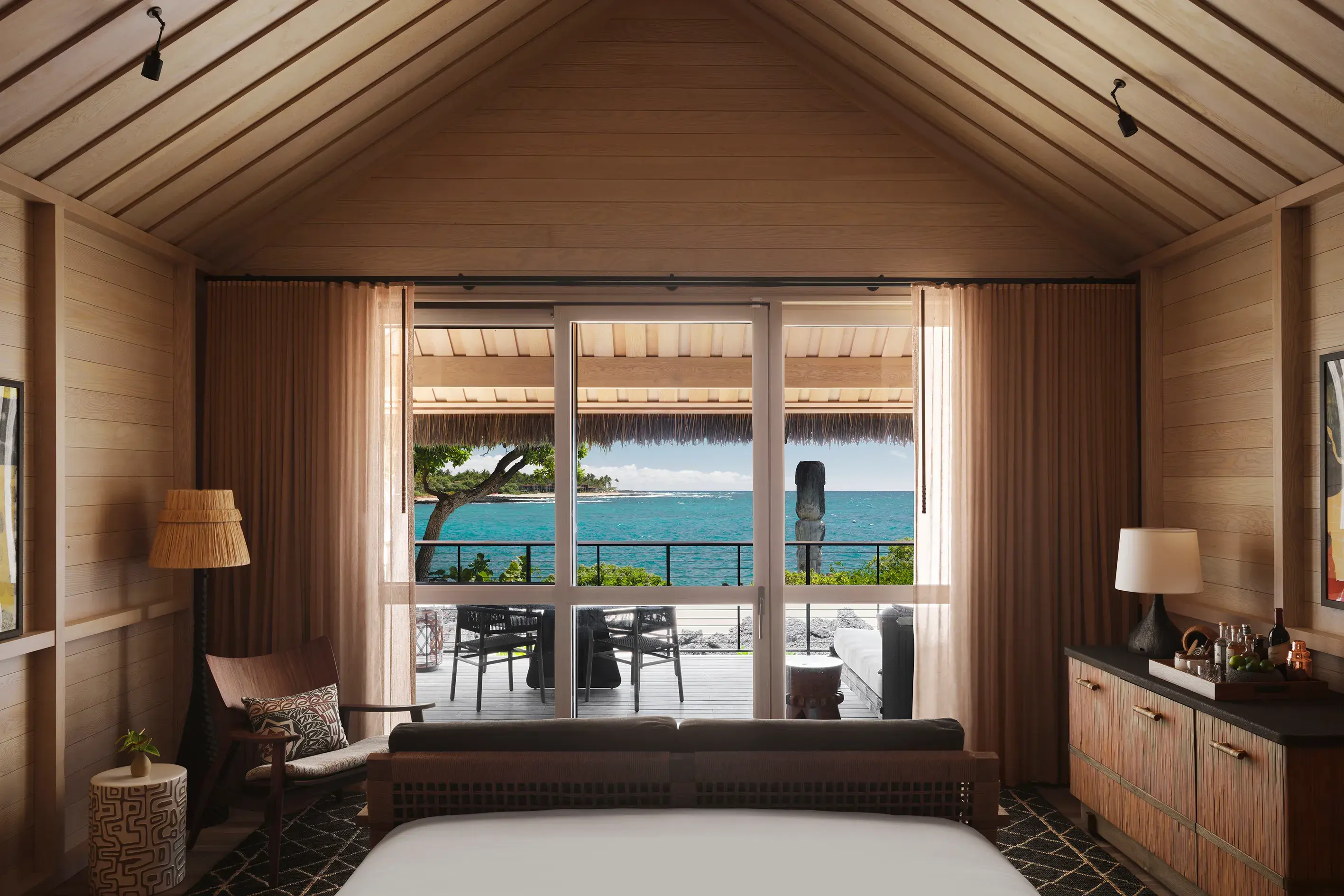 Interior of an Oceanfront Hale at Rosewood Kona Village with deep wood furnishings and large windows looking out over a terrace to the ocean