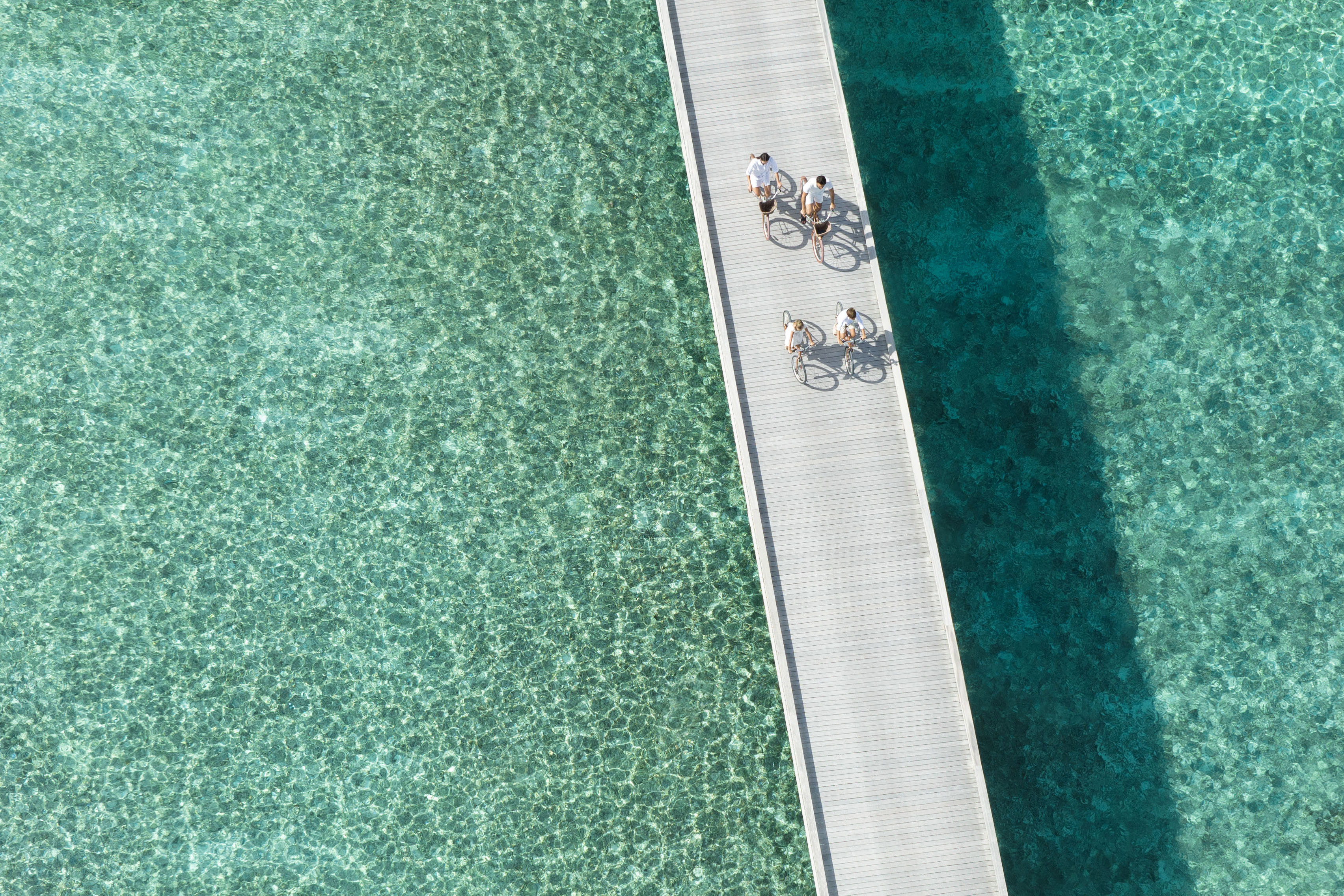 Family of four cycling down a white wood pier over clear blue water