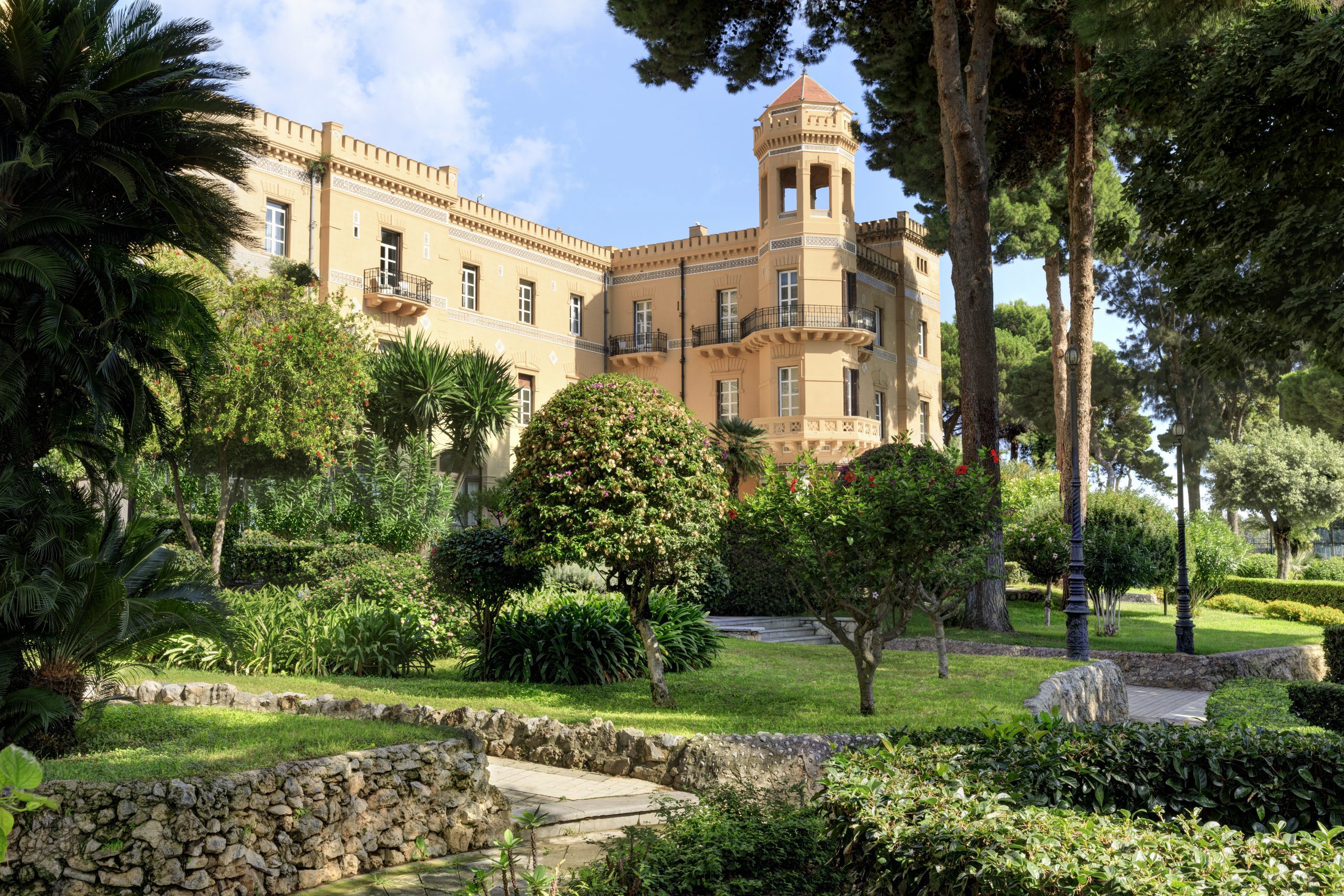 A grand yellow building with a tower, surrounded by lush green gardens and tall trees under a clear blue sky.