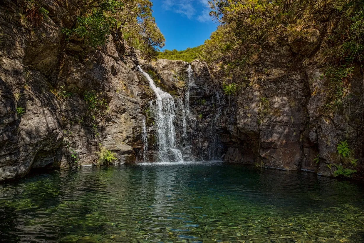 Small waterfall cascading into a clear pool surrounded by rocky cliffs and lush vegetation.