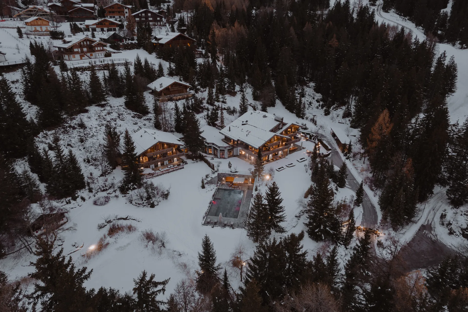 Aerial view of the large snow covered estate surrounding Ultima Crans-Montana featuring two large chalet buildings and pool surrounded by snowy woodland and other buildings