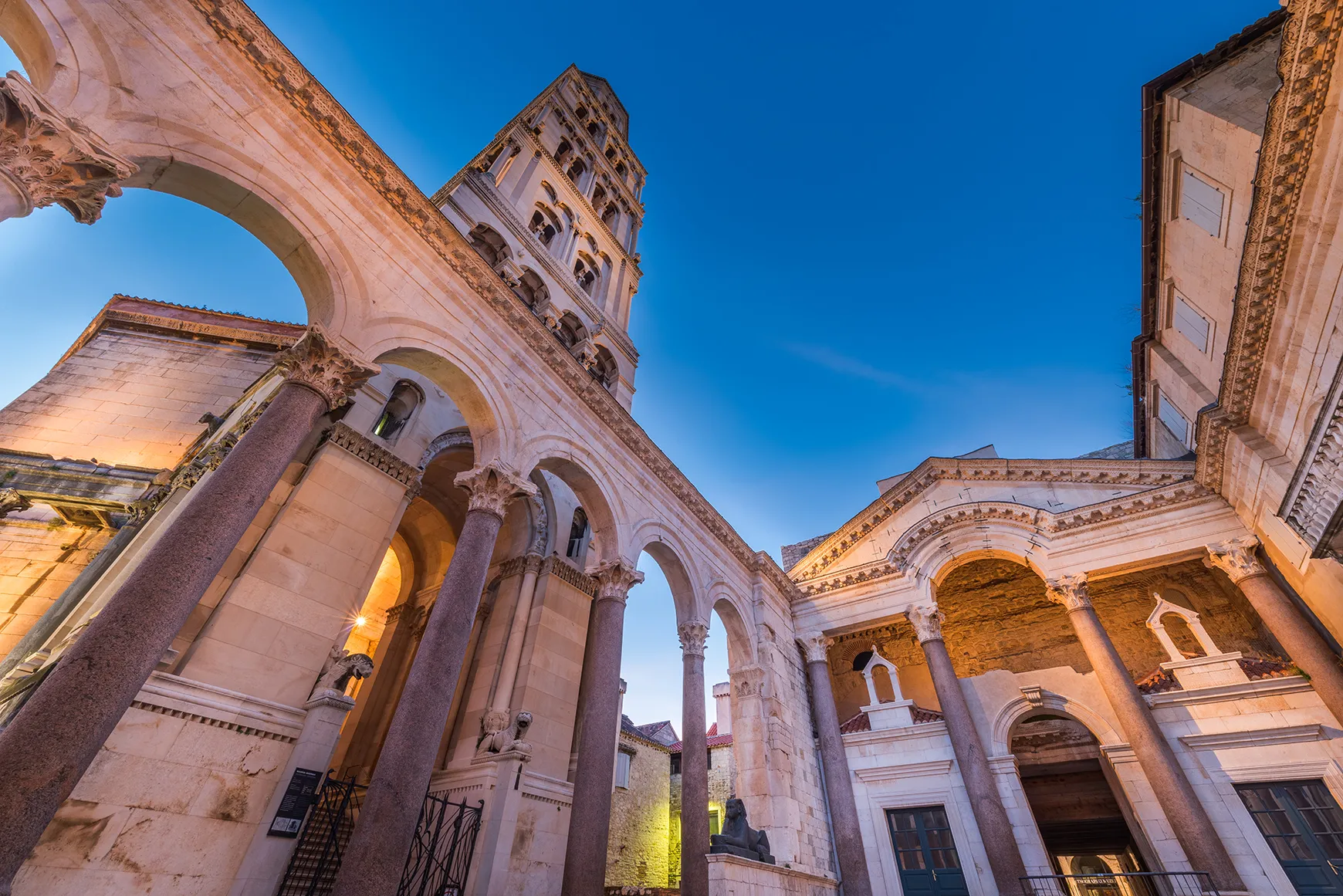 Historic stone courtyard with arched colonnades and a bell tower, viewed against a clear blue sky.