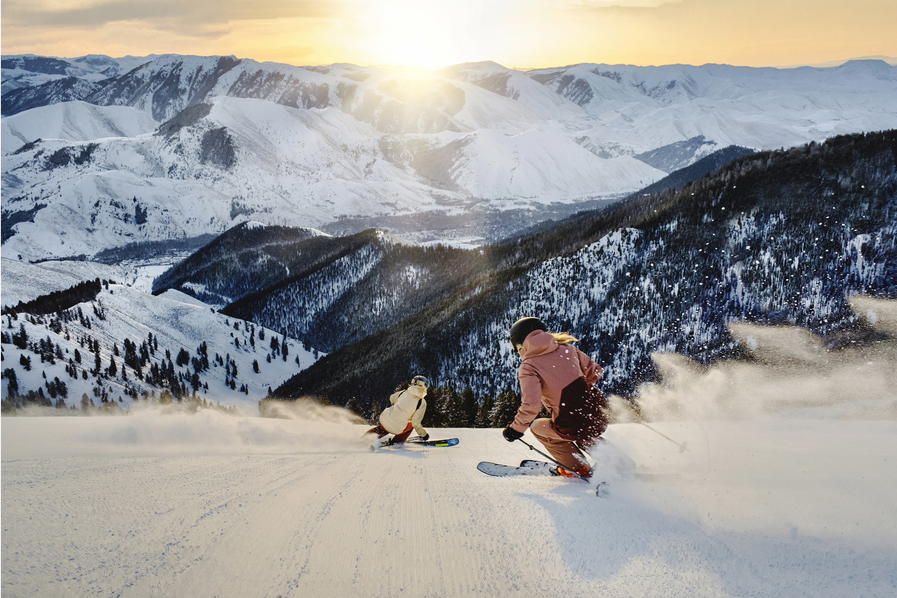 Two skiers descend a snowy mountain slope on Bald Mountain with snow-covered peaks and a setting sun in the background