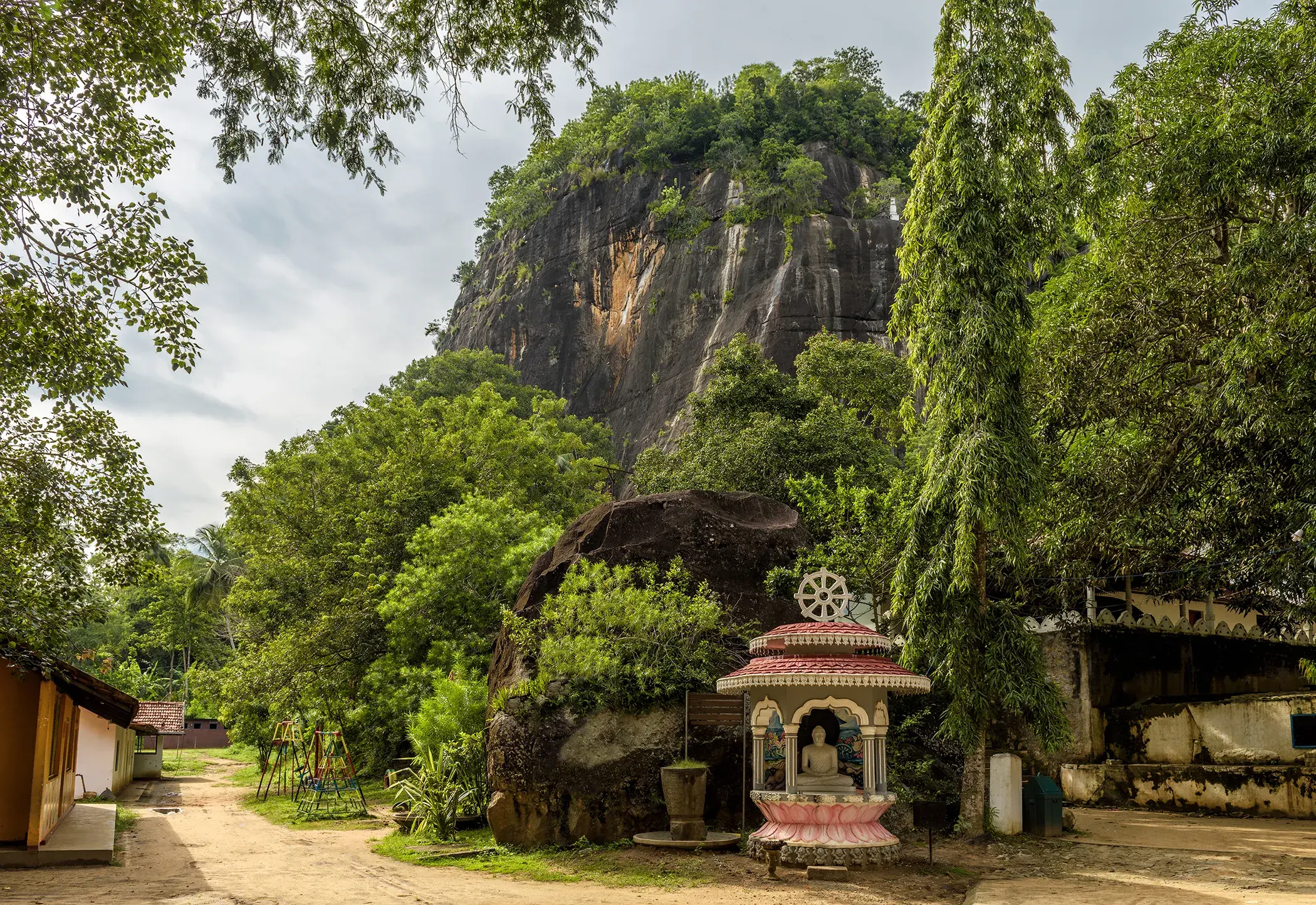 Sacred Buddhist shrine at the temple of Mulgirigala Rock at the base of a lush green rock formation in Sri Lanka, surrounded by tropical trees and natural landscape.