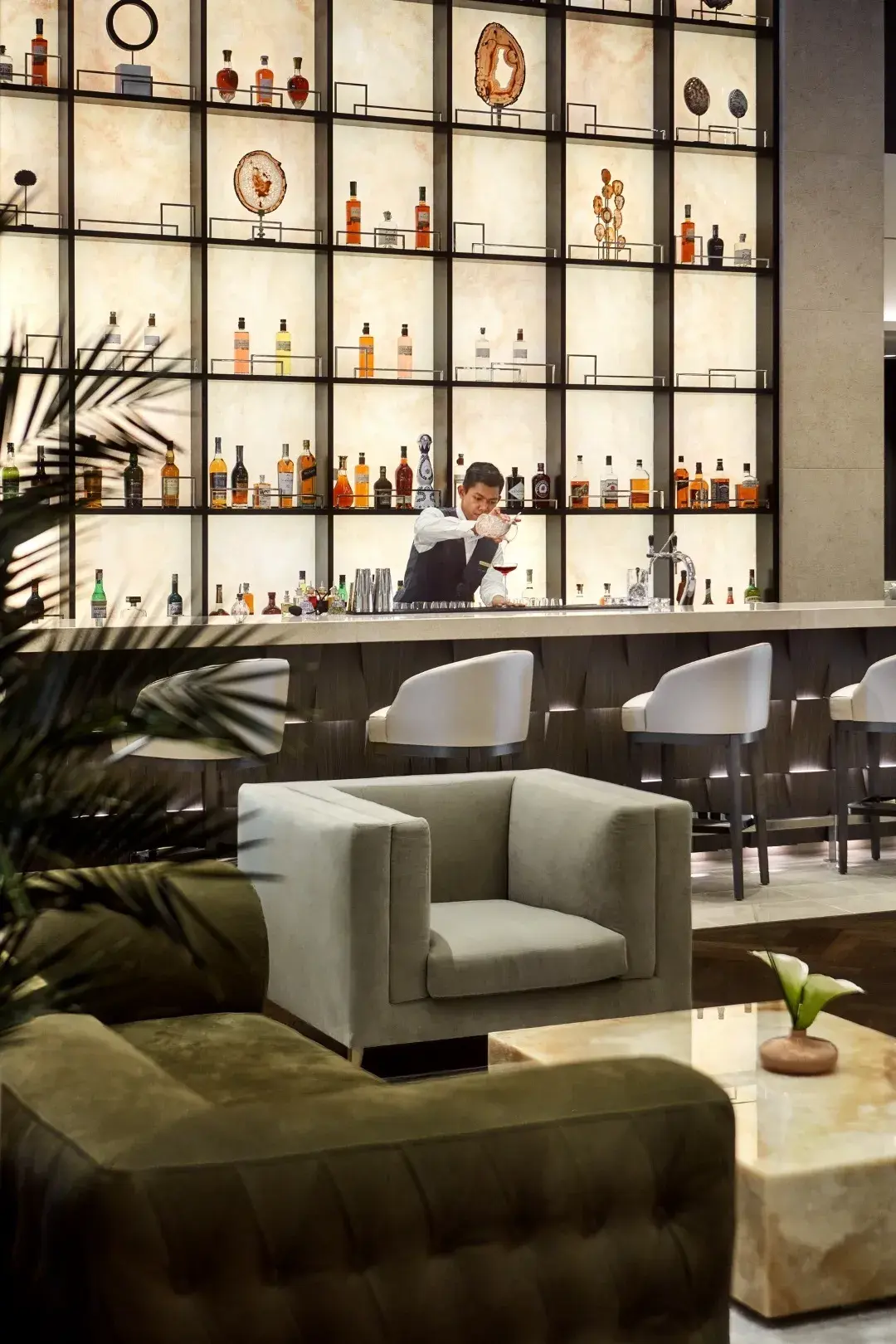 Bartender crafting a drink at a stylish, well-stocked bar with modern seating and decorative shelving.