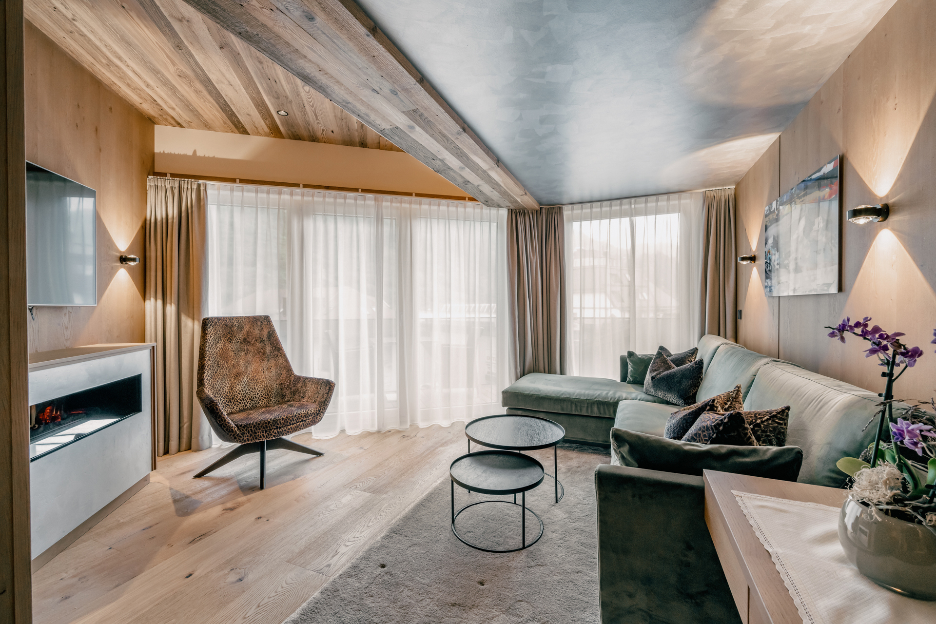 The cozy living room of a Spa Suite at the Gourmet and Wine Hotel Austria with an L-shaped sofa, modern armchair, round coffee tables, and floor-to-ceiling windows with sheer curtains
