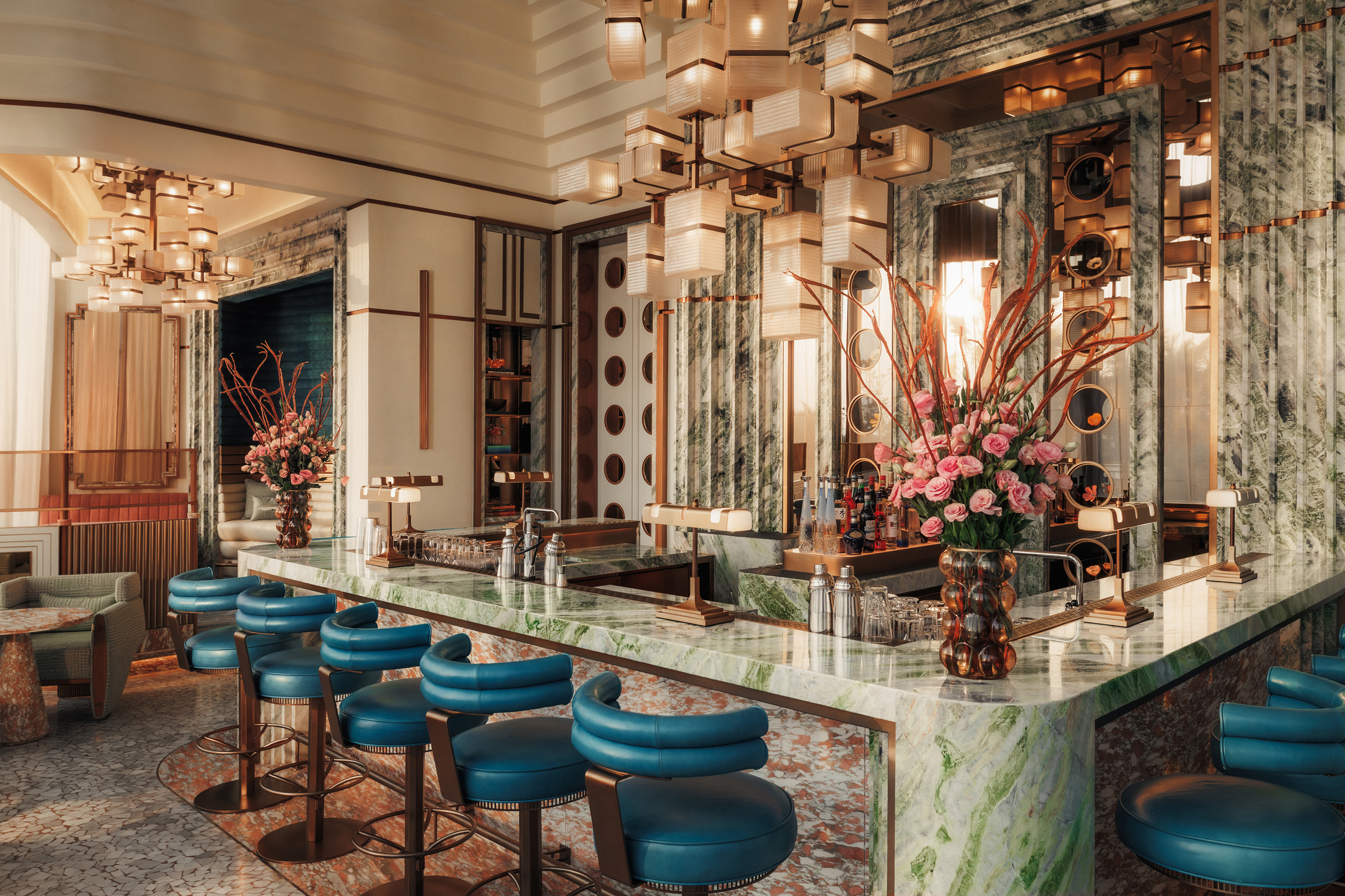 An ornate marble bar in the Italian Bar at Jumeirah Marsa Al Arab surrounded by blue bar stools beneath a statement chandelier with glimpses of other furniture around