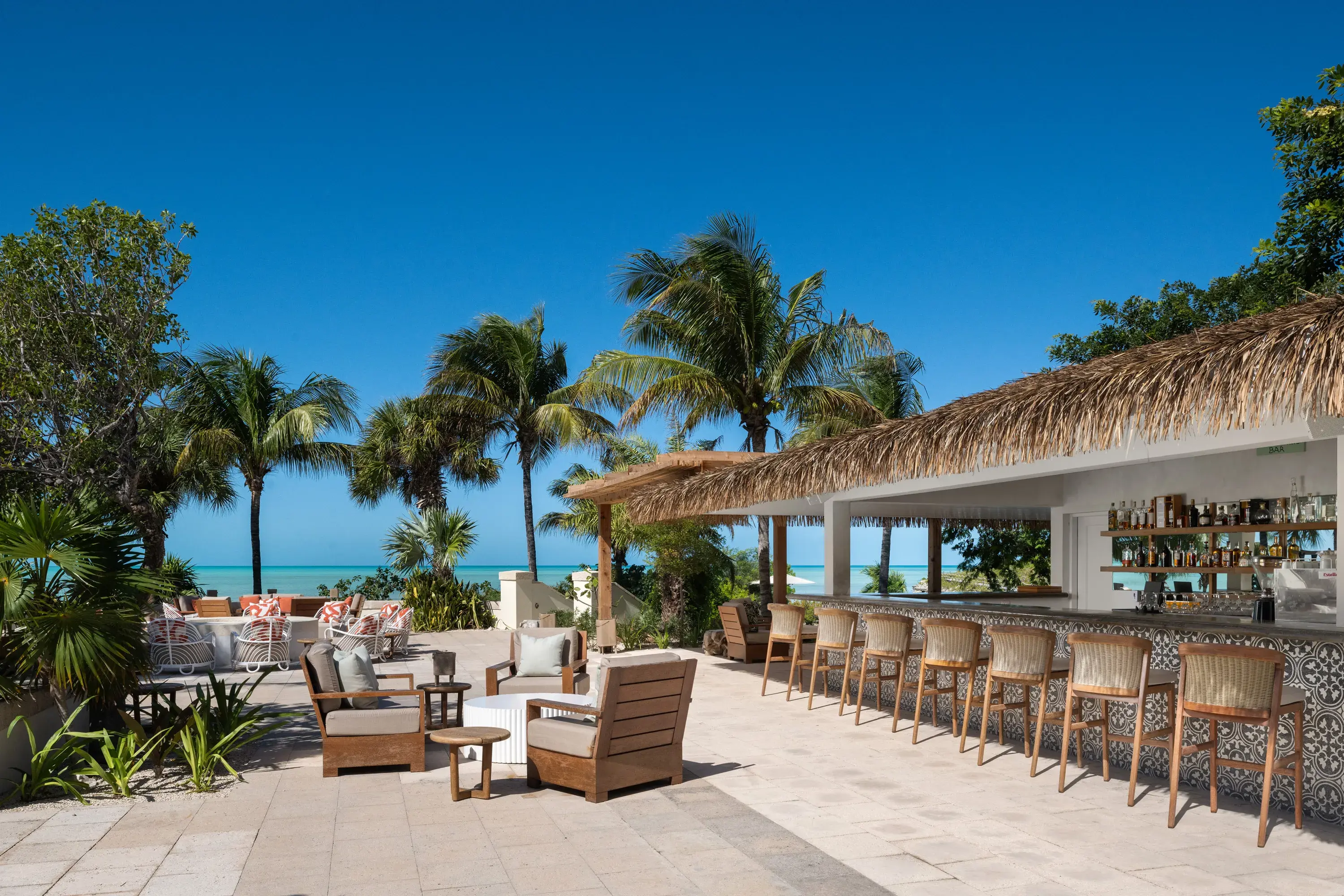 Bar area of Calico Mahoneys Bar at Ambergris Cay in Turks & Caicos featuring a thatched bar area lined with chairs and comfy lounge chairs surrounded by palms beneath a clear blue sky