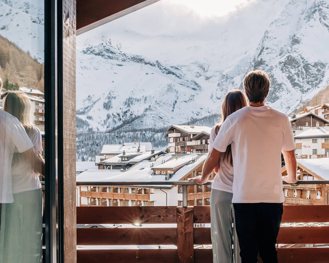 Two guests standing on a balcony overlooking a snowy alpine village with dramatic mountain views.