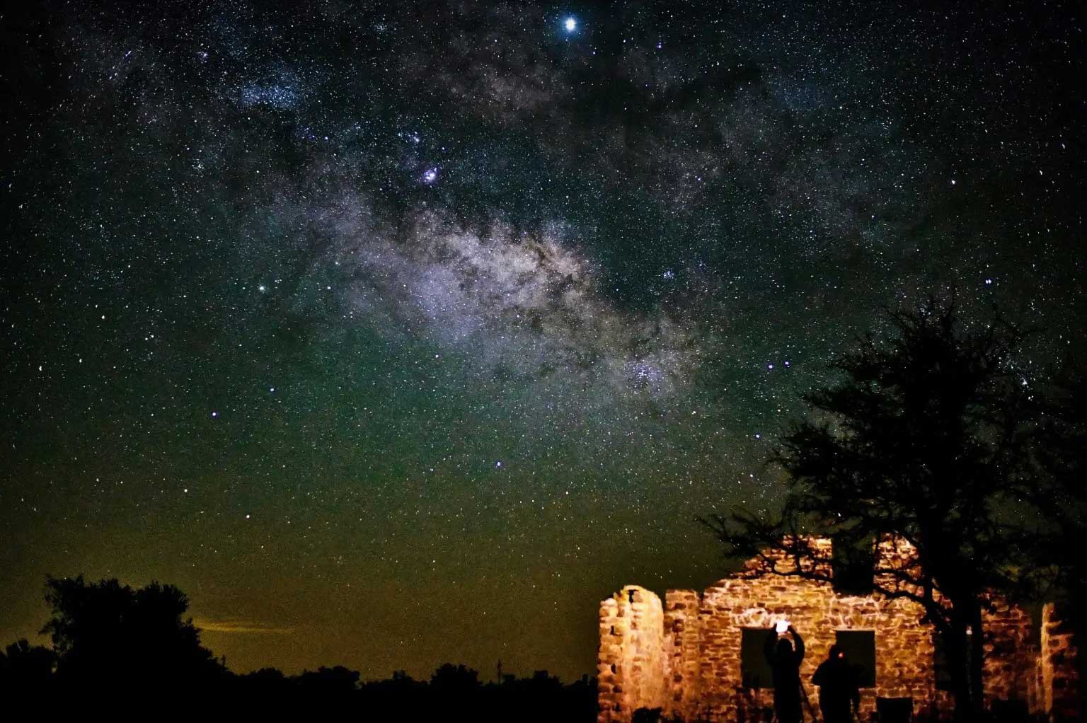 Star-filled night sky with the Milky Way visible above a rustic stone building and silhouetted figures.