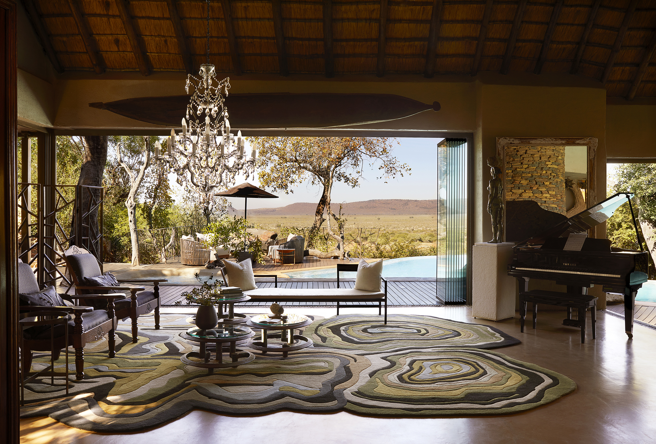An ornate safari-chic lounge featuring a chandelier, grand piano and furniture with wide sliding doors leading to the pool opened up to reveal a sweeping view of the landscape 