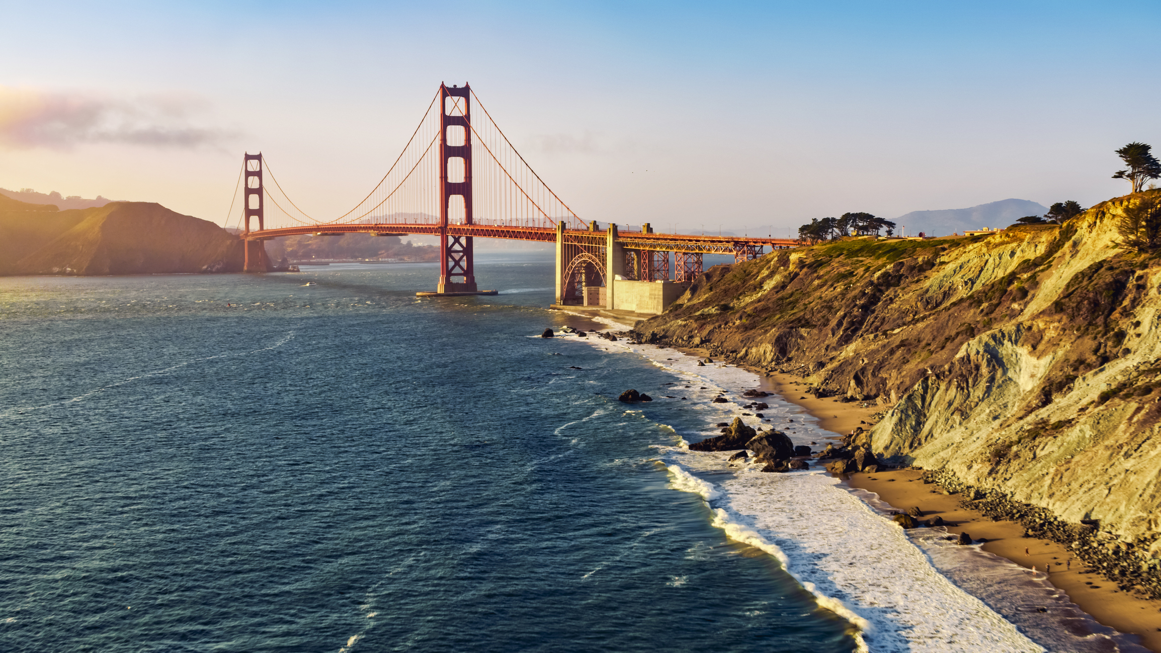 The Golden Gate Bridge during late afternoon with warm sunlight and a partly cloudy sky, spanning across a body of water with a rocky shoreline in the foreground.