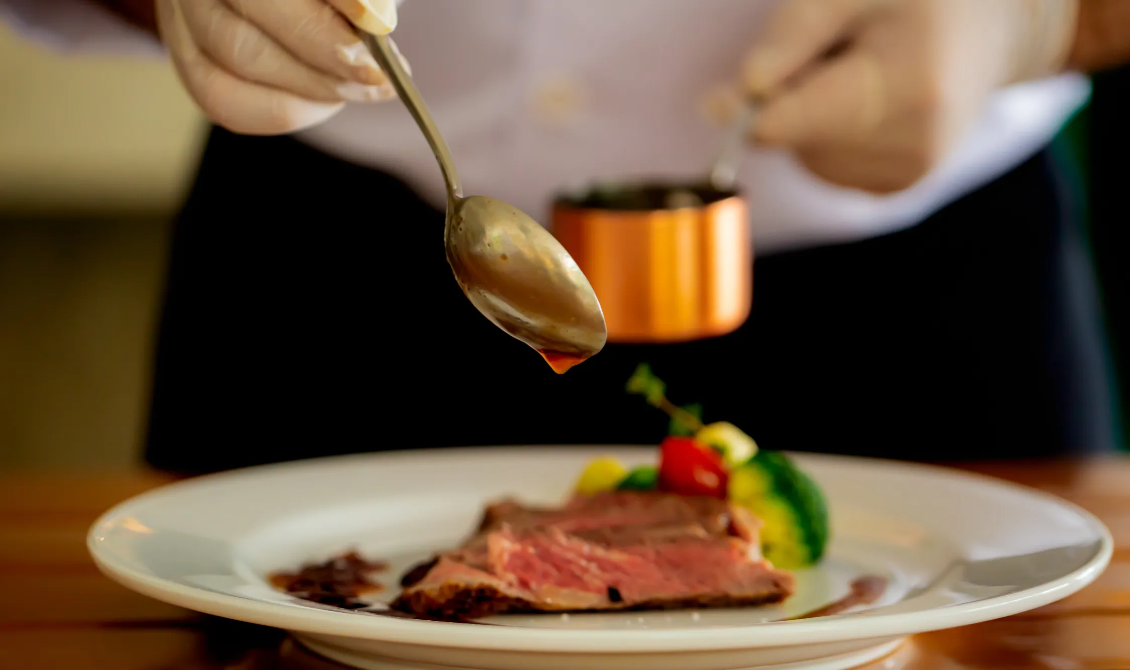 A chef pours sauce over cooked meat on a white plate with vegetables including broccoli and cherry tomatoes.