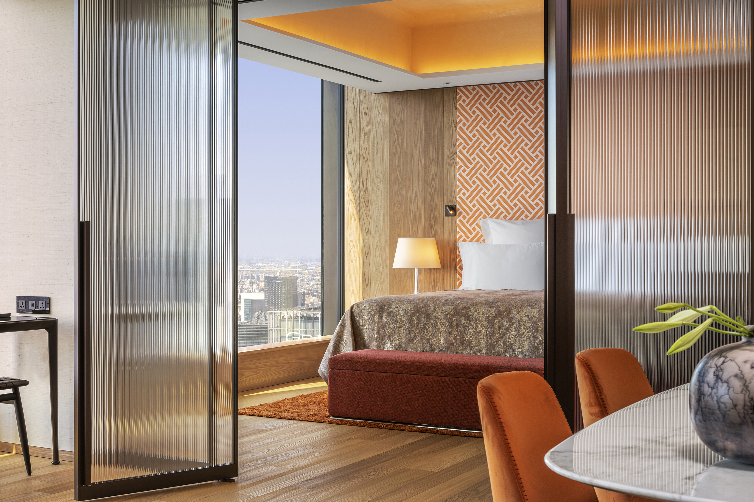 A partial view of a suite bedroom with comfy bed and side lamp through a ribbed glass wall with a partial view of a table and vase on the other side