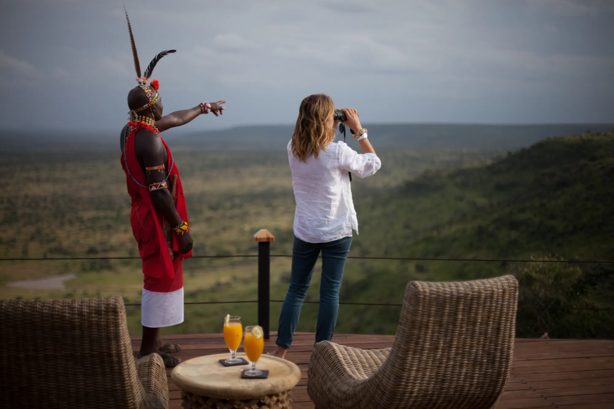 Two people on the observation deck at Elewana Loisaba Tented Camp with one person in traditional Maasai dress pointing out something in the distance to the other person looking through binoculars 