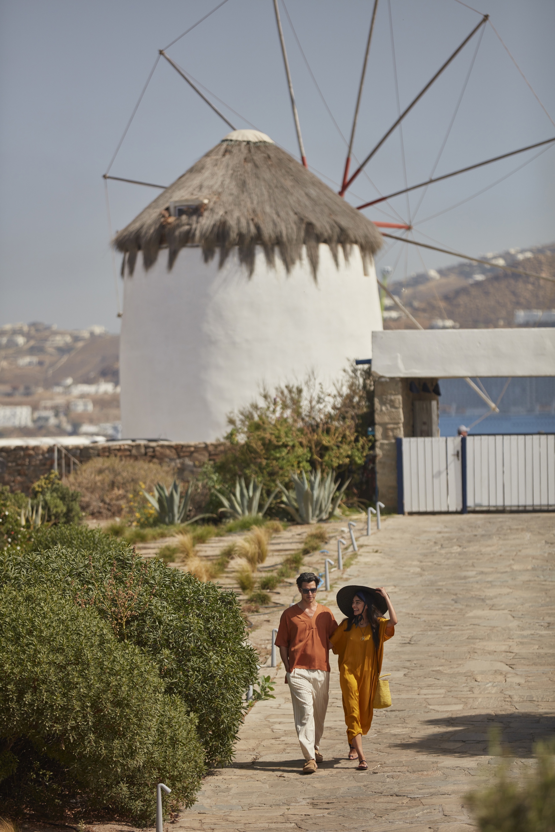 A couple dressed in summery clothing walking away from one of the iconic white windmills on Mykonos beside greenery
